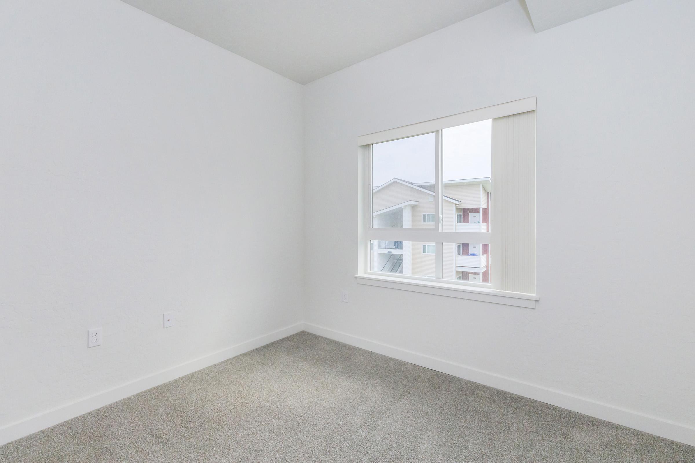 A bright, empty room with light gray carpeting and white walls. A large window allows natural light to enter, revealing views of the outside. The simplicity of the decor creates a minimalist atmosphere.