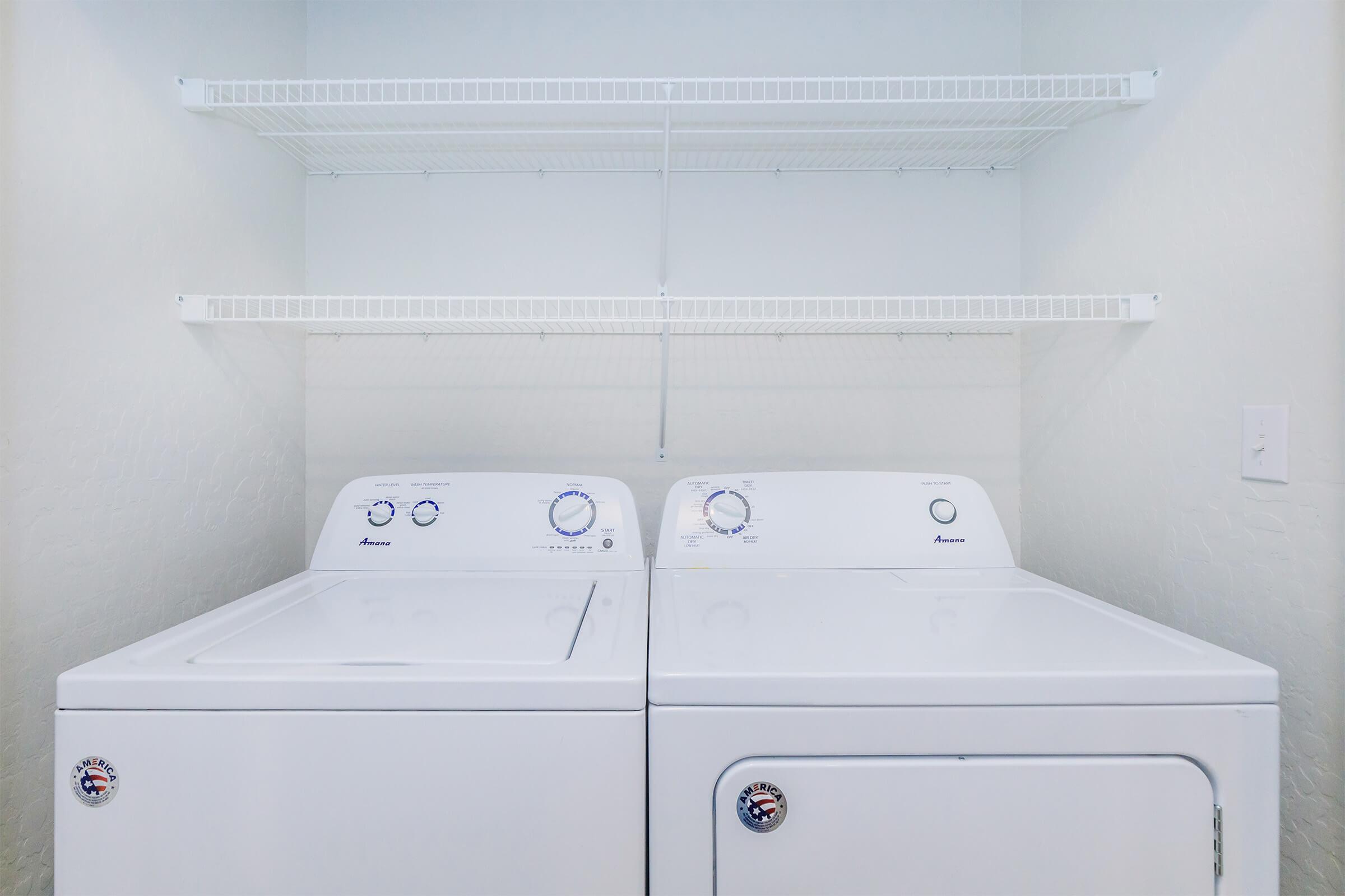 A clean laundry room featuring a washing machine and a dryer side by side. Above them, there are white wire shelves for storage. The walls are painted a light color, creating a bright and uncluttered space, ideal for laundry tasks.