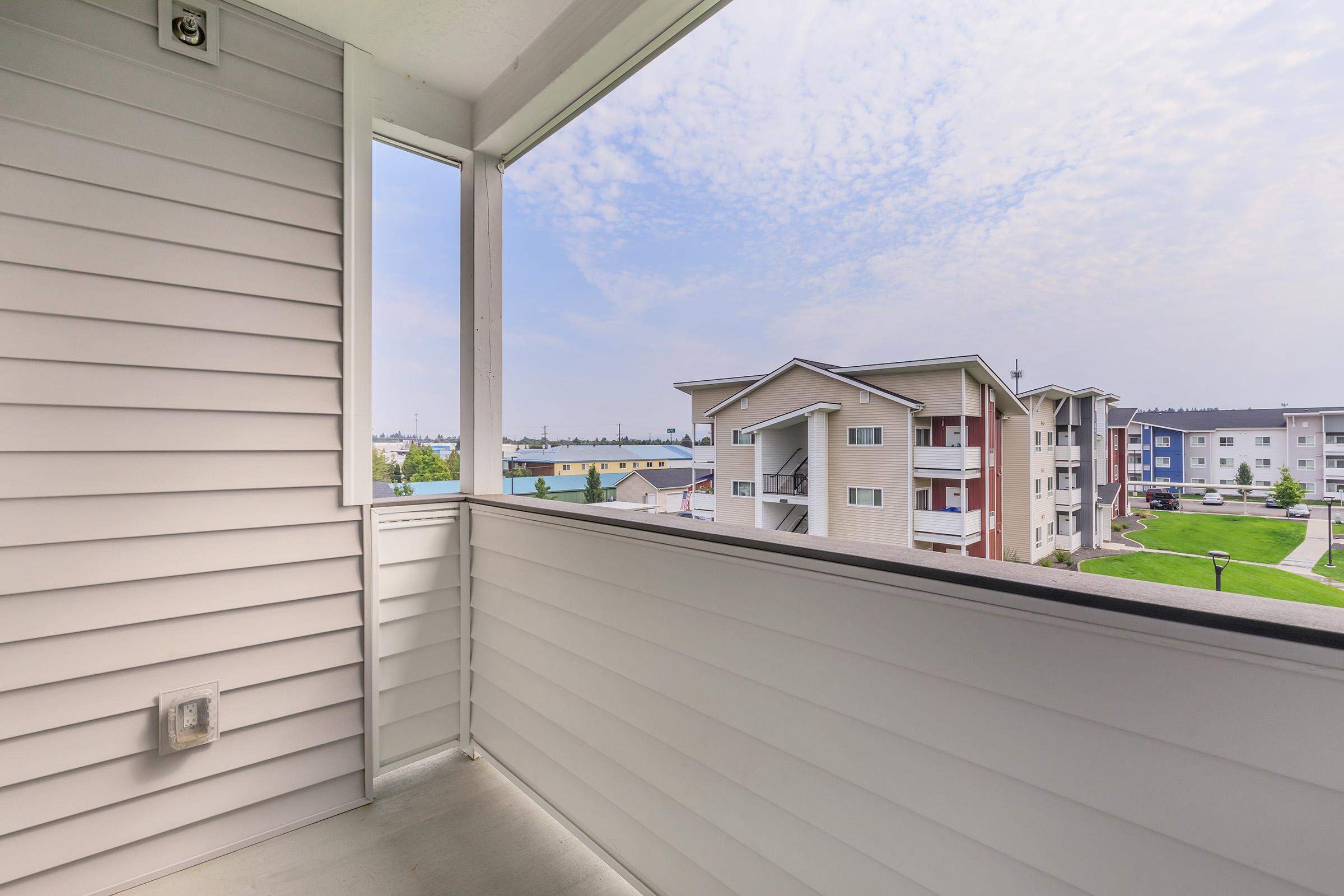 A view from a balcony showing white siding and a clear sky. Nearby buildings with multi-colored exteriors can be seen in the background, along with green landscaping and outdoor lighting fixtures. The scene conveys a peaceful residential area.