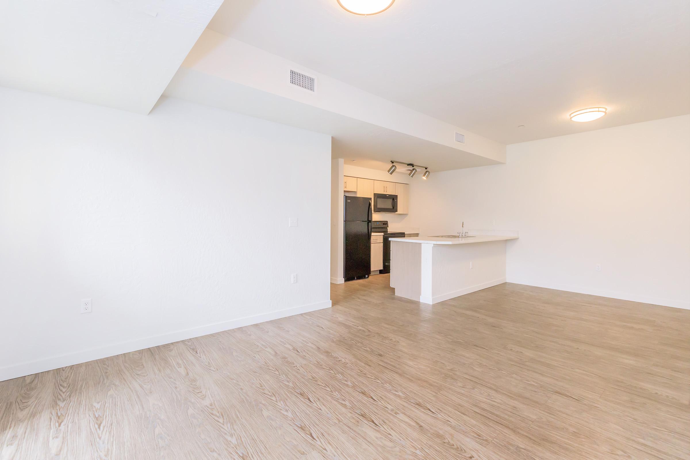 Spacious, modern apartment interior with light wood flooring and white walls. A kitchen area is visible in the background, featuring dark cabinets and stainless steel appliances. Natural light enters through the open space, creating a bright and inviting atmosphere.
