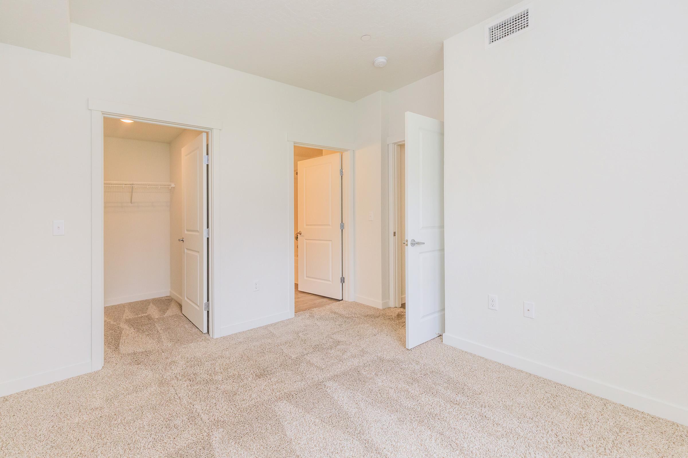A spacious, well-lit room featuring light-colored walls and plush carpet. Two doors lead to adjacent areas, one likely a closet. The room has a clean and modern feel, with natural light streaming in from an unseen source. Ideal for a bedroom or office space.