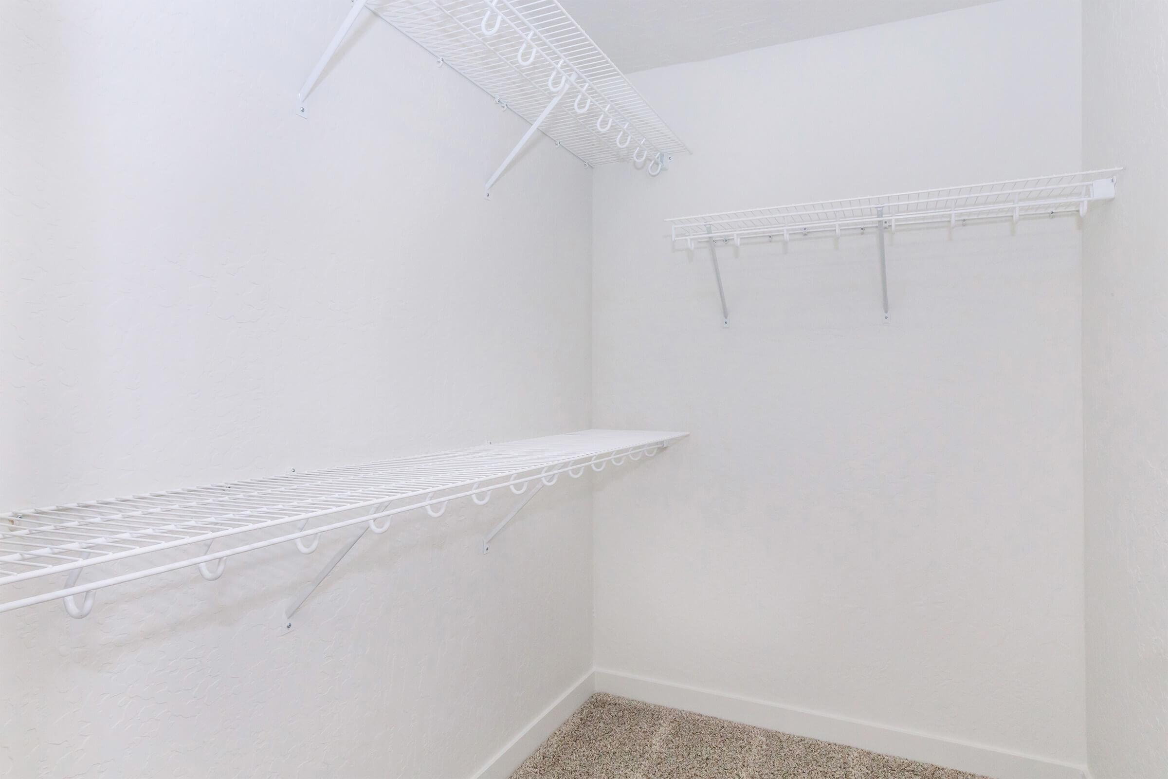 A clean, empty closet featuring white wire shelving. The walls are painted white, and the floor is covered with a light-colored carpet. There are two shelves: one long shelf on the back wall and a shorter shelf mounted higher on the side wall, both providing storage options.