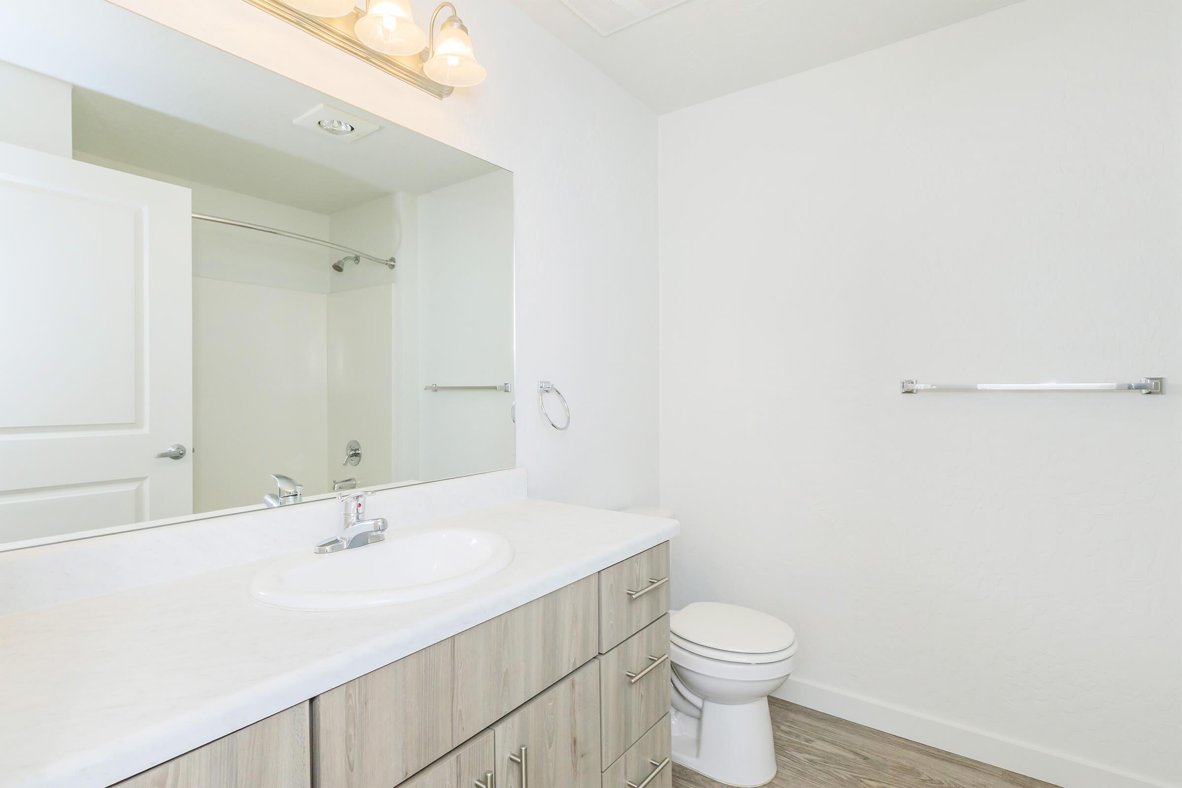 A clean, modern bathroom featuring a large mirror above a white sink with a marble countertop, a wooden vanity with multiple drawers, a toilet, and a shower area in the background. Soft lighting from above enhances the bright, neutral color scheme of the room.