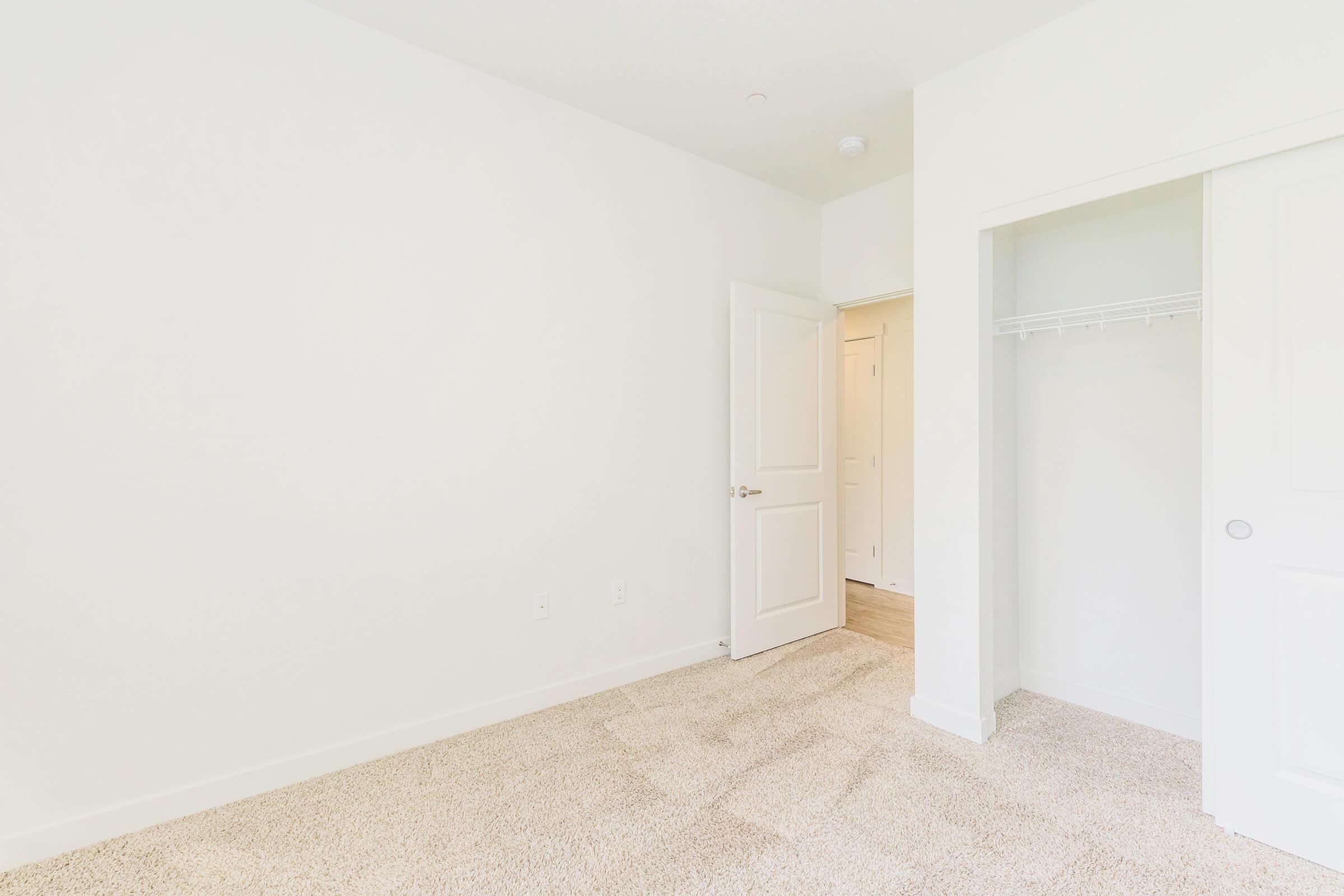A bright, empty room with white walls and carpet. There is a closet with sliding doors on the left and a partially open door leading to another area on the right. The space is well-lit, creating a clean and spacious atmosphere.