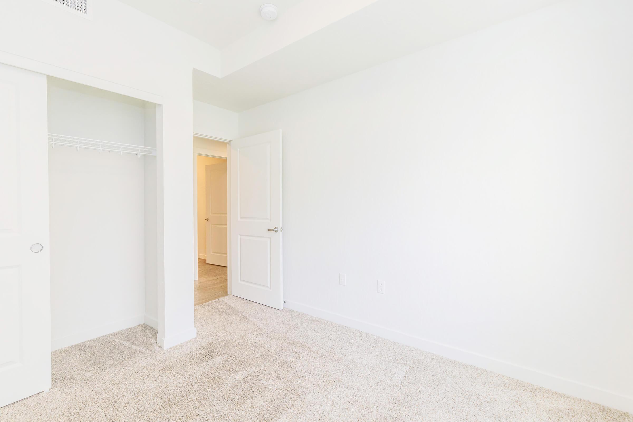 A bright, empty room featuring light-colored walls and beige carpet. There is a closet with sliding doors on the left and an open doorway leading to another room. The overall atmosphere is clean and spacious, with plenty of natural light.