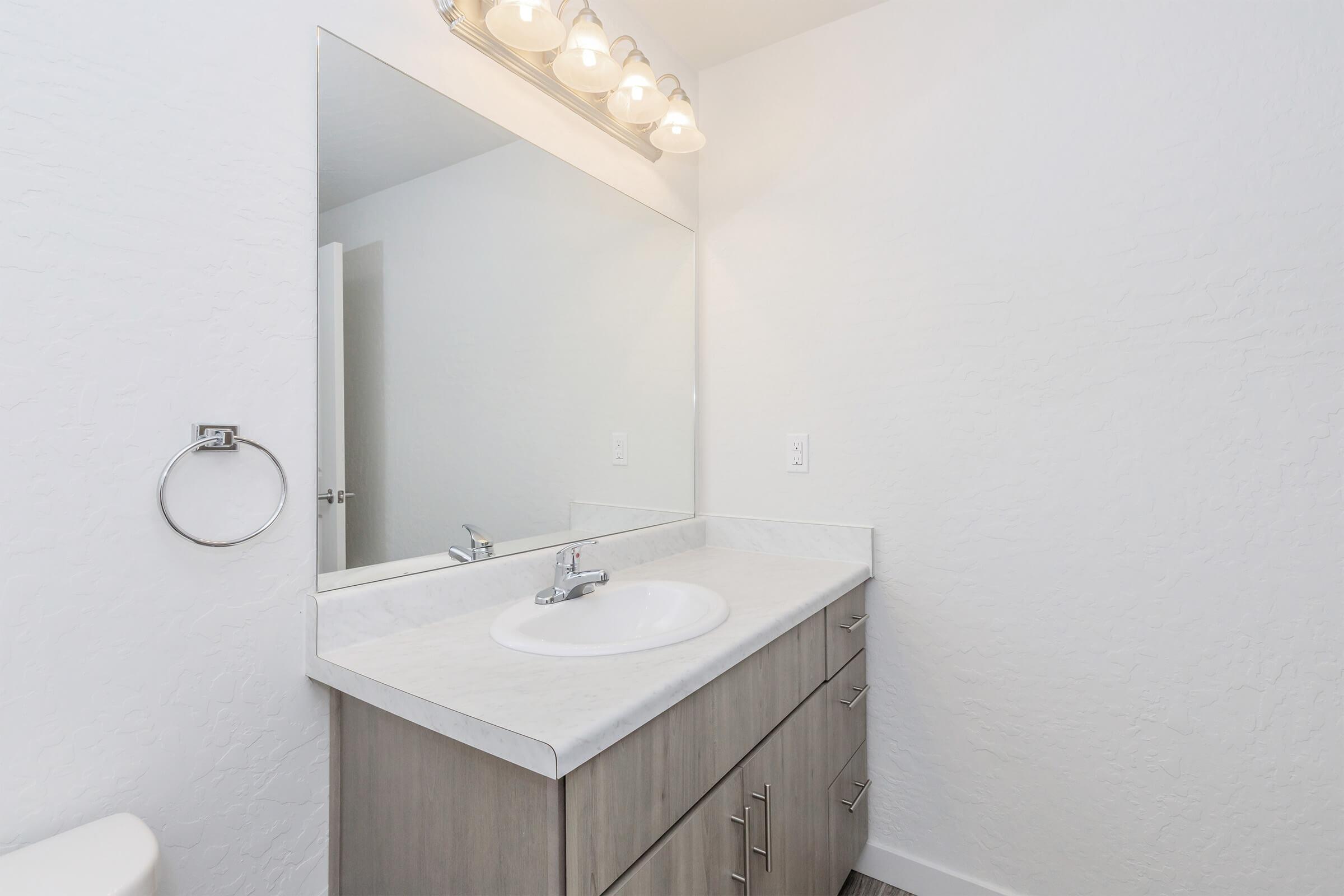 Modern bathroom interior featuring a simple vanity with a white countertop and a single sink, complemented by a large mirror above. The walls are painted white, and the lighting is provided by four overhead light fixtures. A towel ring is mounted near the sink, contributing to a clean and minimalist aesthetic.