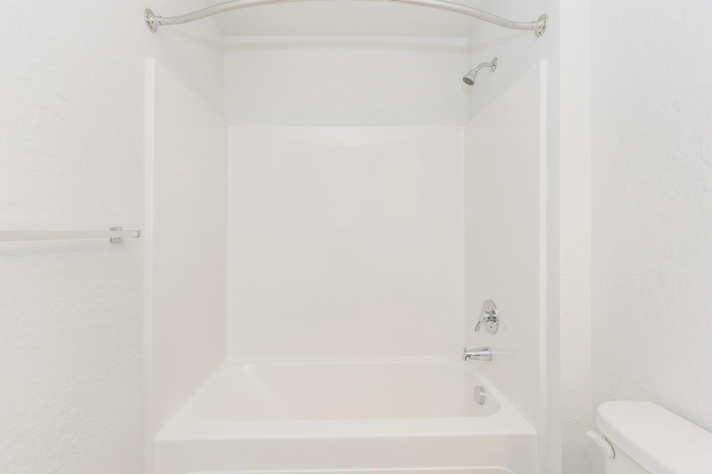A clean and minimalistic bathroom featuring a white bathtub with an attached showerhead. The walls are plain white, and there is a modern faucet on the side of the bathtub. A towel bar is mounted on the wall, and the space appears bright and uncluttered.