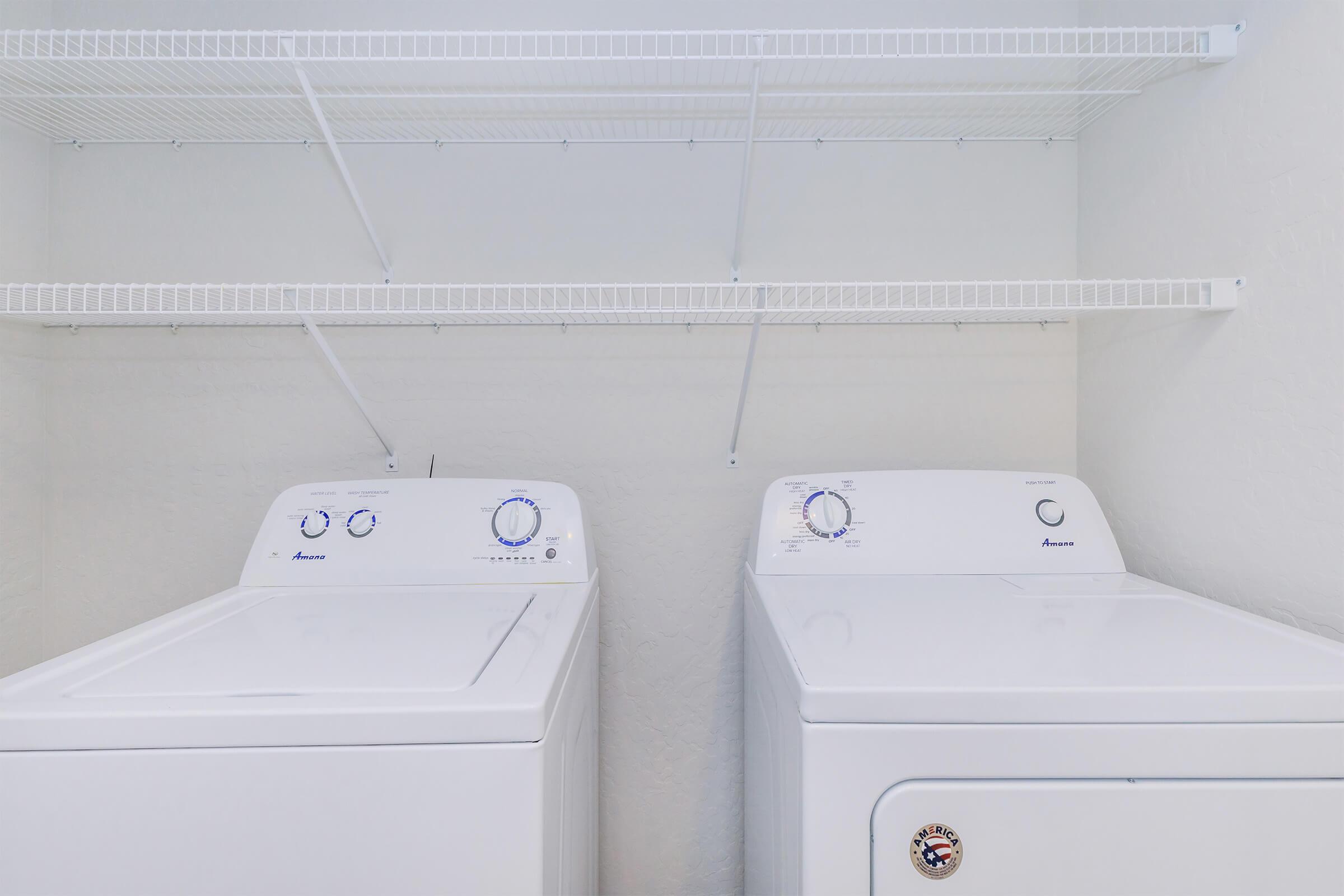 A clean laundry room featuring a white washing machine and a matching dryer side by side. Above them, there are two wire shelves for storage. The walls are painted white, creating a bright and tidy atmosphere.