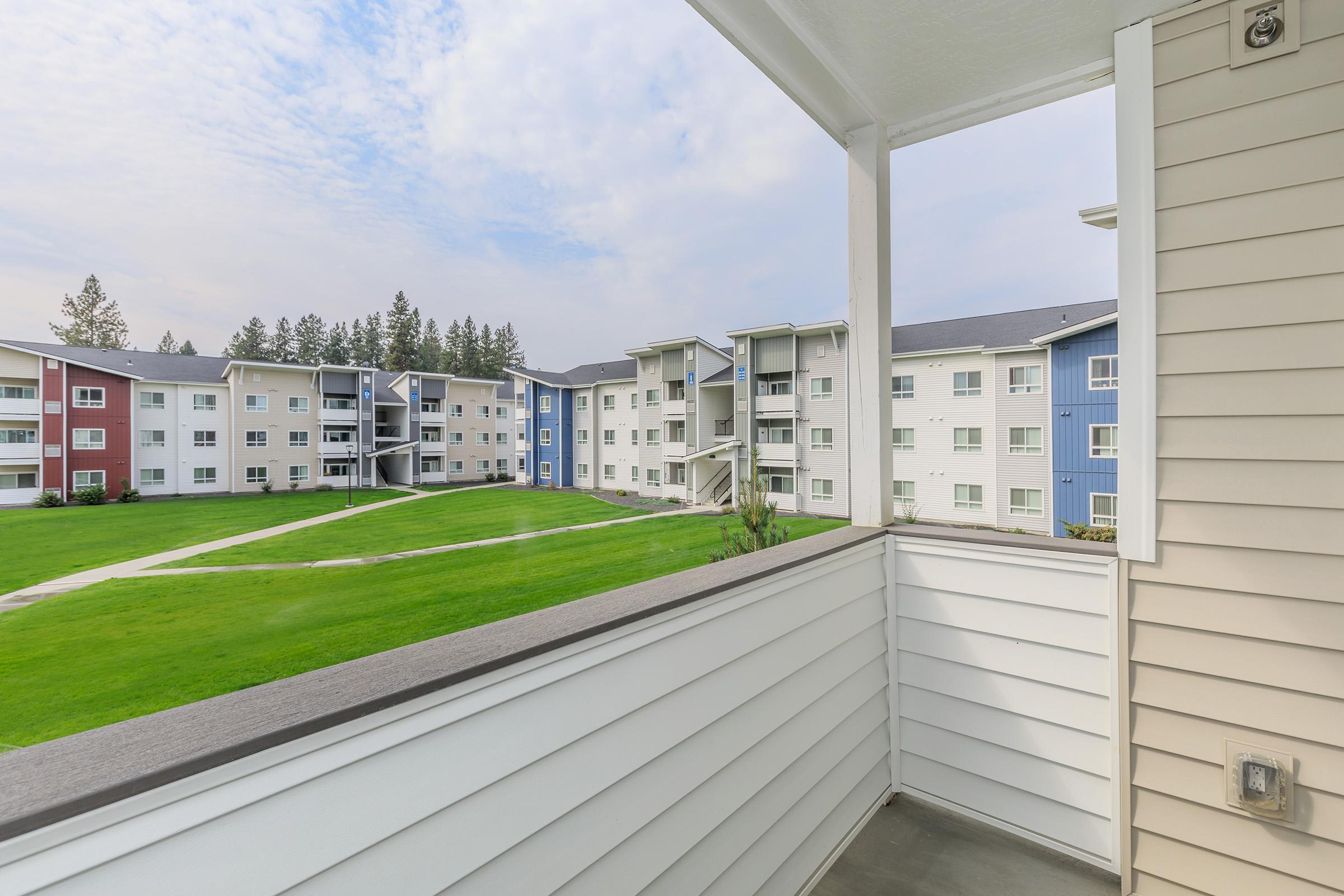 View from a balcony overlooking a landscaped courtyard with green grass and a pathway, surrounded by multi-story apartment buildings featuring a mix of white, blue, and red exteriors under a cloudy sky.