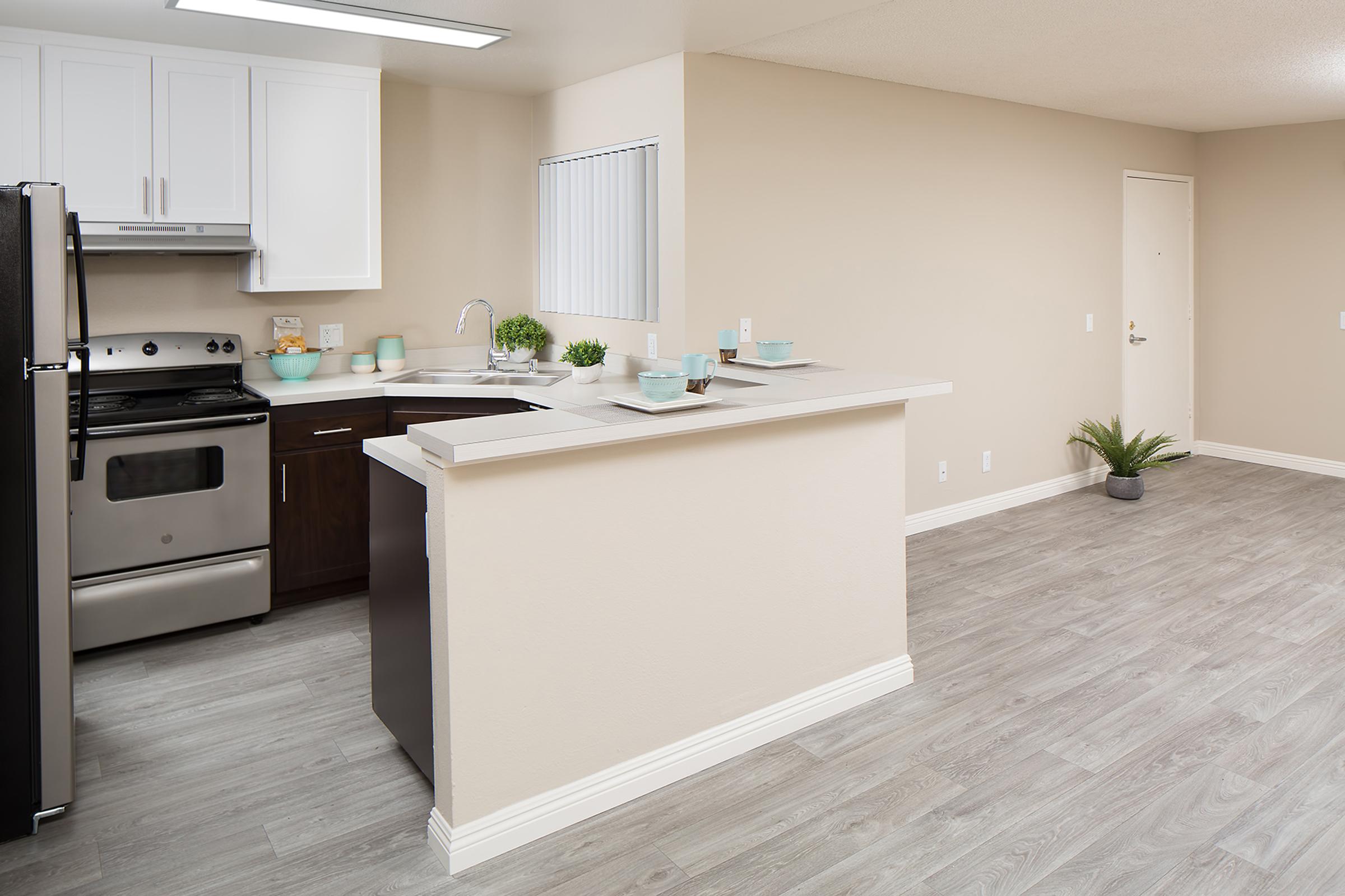 A modern kitchen and living area featuring an open layout. The kitchen has black appliances, white cabinets, and a countertop with light blue dishes. The living space has light wood flooring and pale beige walls, with a small potted plant on the countertop and near the entrance.