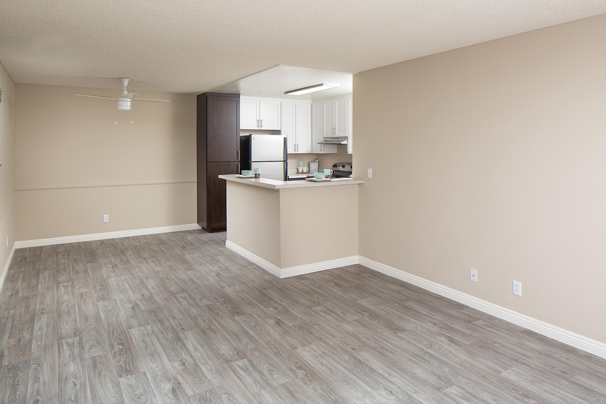 A spacious, empty living area featuring light-colored walls and gray wood-like flooring. To the left, a ceiling fan is visible. An open kitchen with white cabinetry, a stainless steel refrigerator, and a countertop bar area is seen in the background, creating a modern and airy atmosphere.