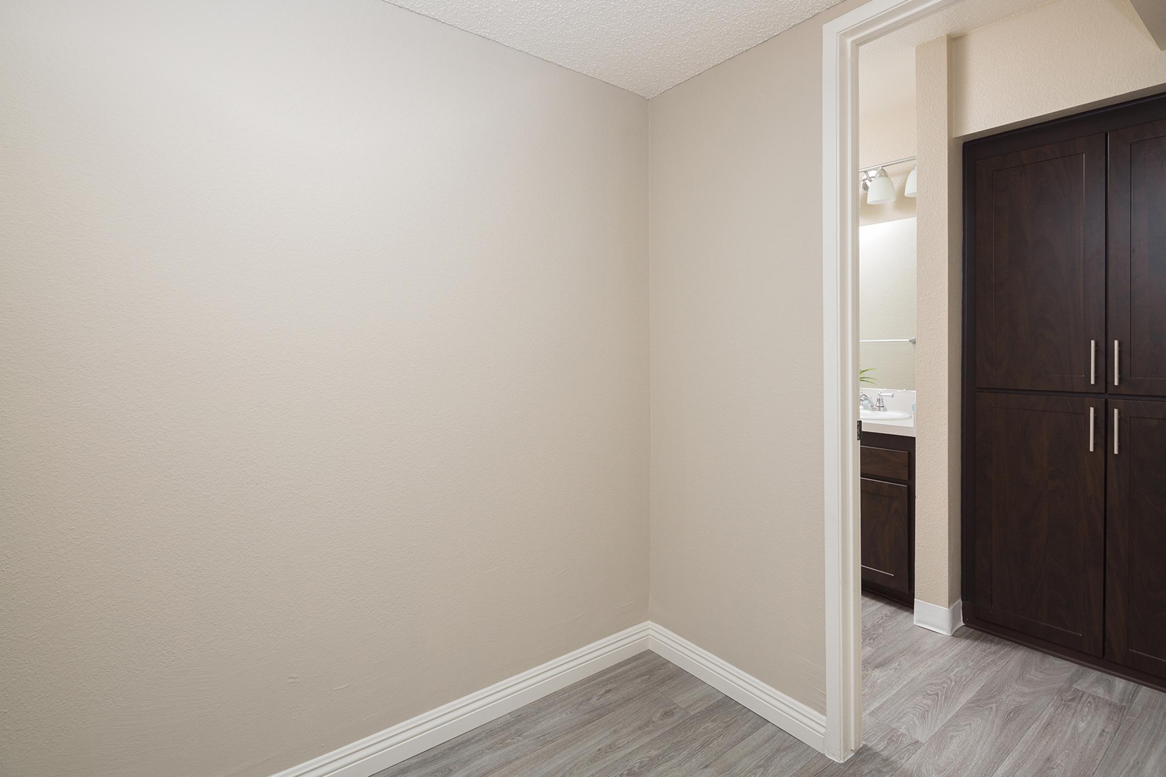 A simple interior view of a bathroom area showing a blank beige wall and a doorway leading to a bathroom with dark cabinetry visible. The flooring is light gray. Natural light comes from the bathroom area, enhancing the neutral tones of the space.