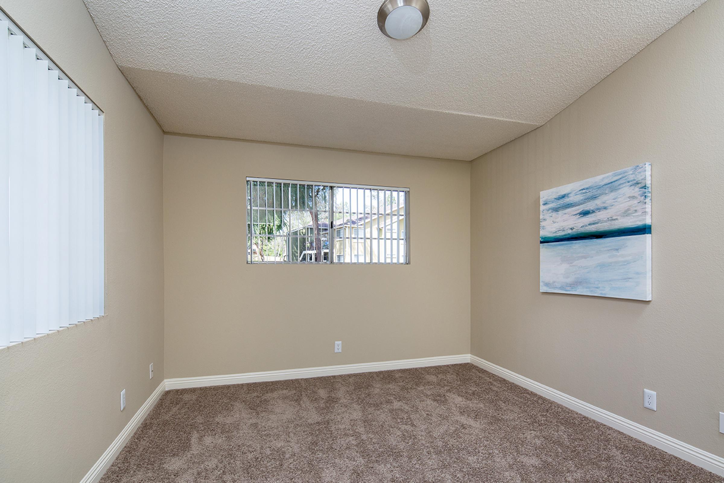 A small interior room featuring light beige walls and gray carpet. There is a window with vertical blinds on one wall, allowing natural light. Another wall displays a framed abstract painting with blue and white colors. The ceiling has a simple light fixture, adding to the room's minimalist decor.