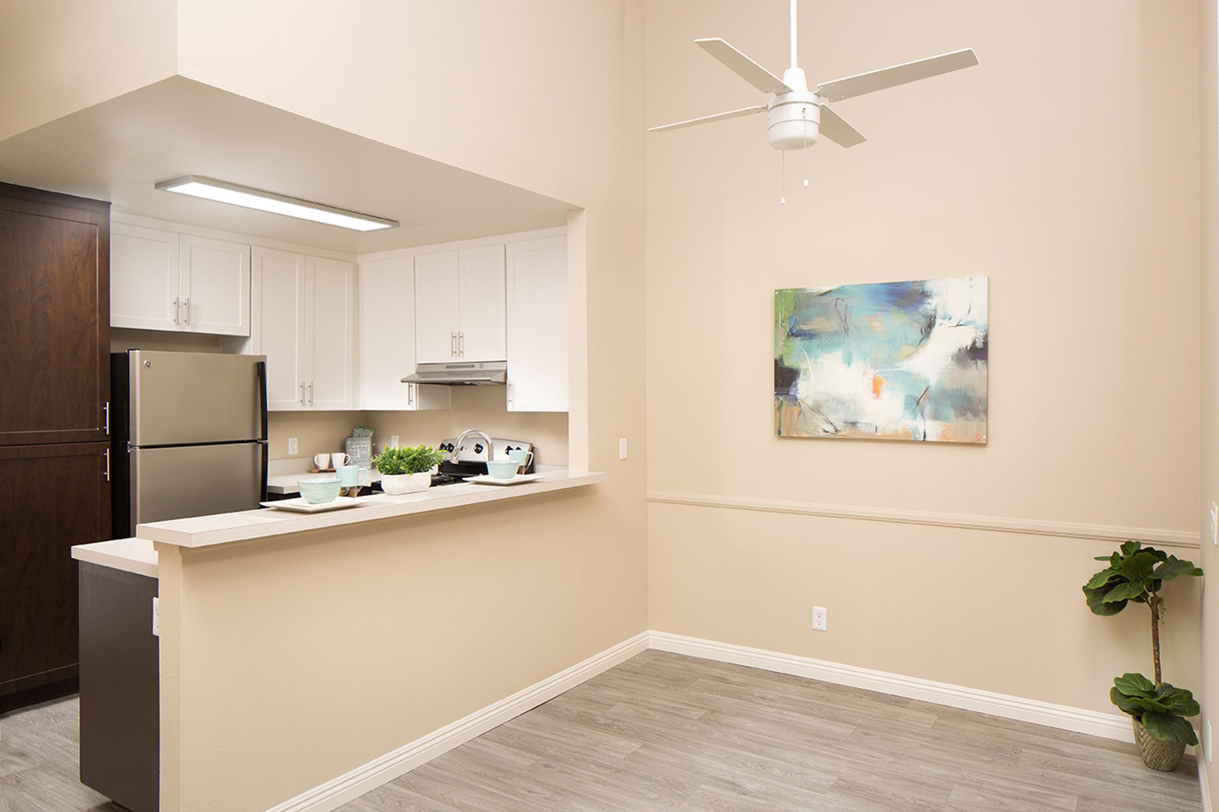 A modern kitchen space featuring white cabinetry, stainless steel appliances, and a light wood floor. A small dining area is visible with a decorative plant and an abstract painting on the wall, complemented by a ceiling fan for ventilation. The walls are painted in a soft beige.
