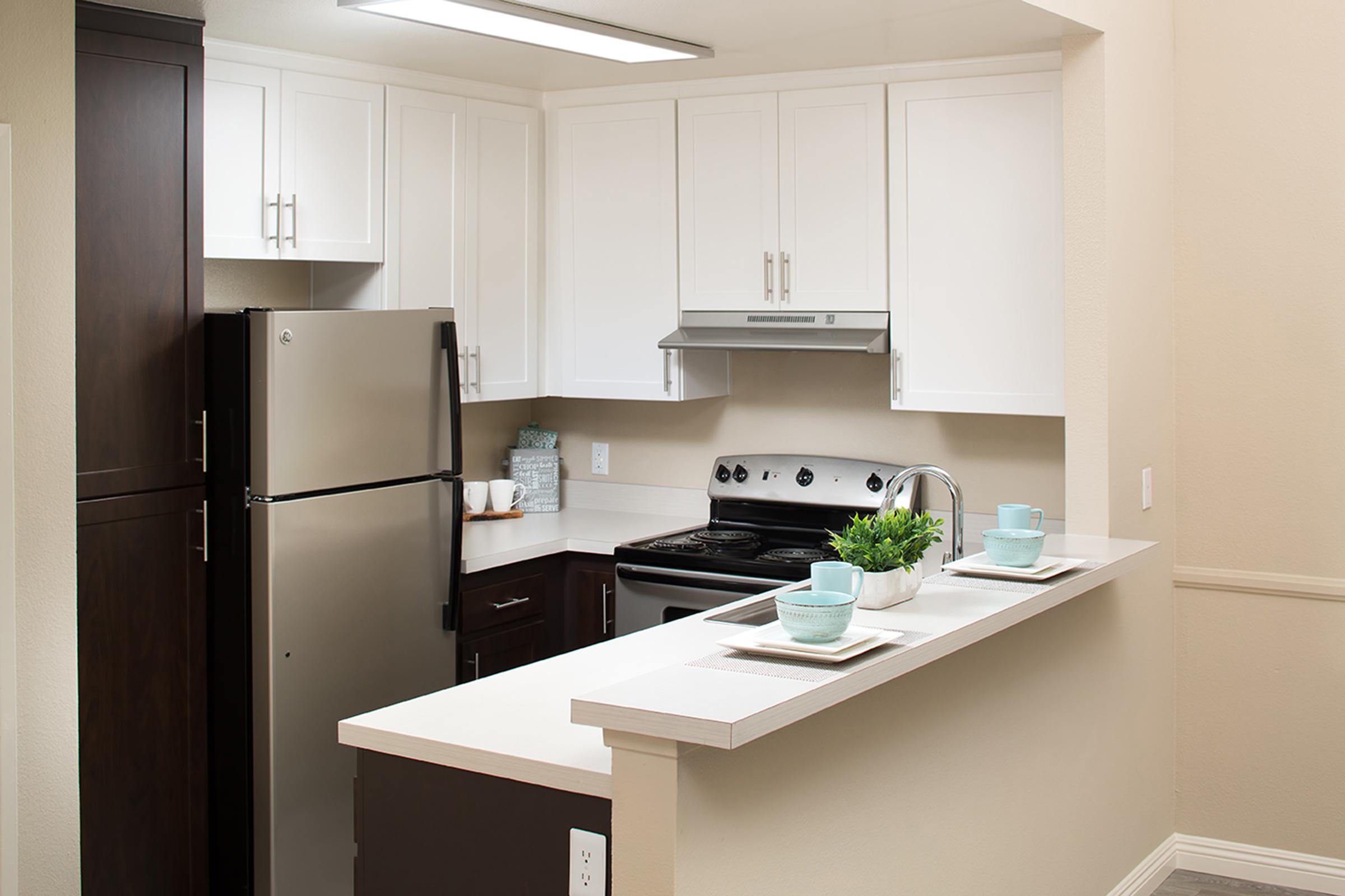 A modern kitchen featuring stainless steel appliances, including a refrigerator and stove, with white cabinets and a light countertop. The kitchen has a bar-style layout with two teal cups on a tray and a small plant as decor, creating a clean and inviting atmosphere.