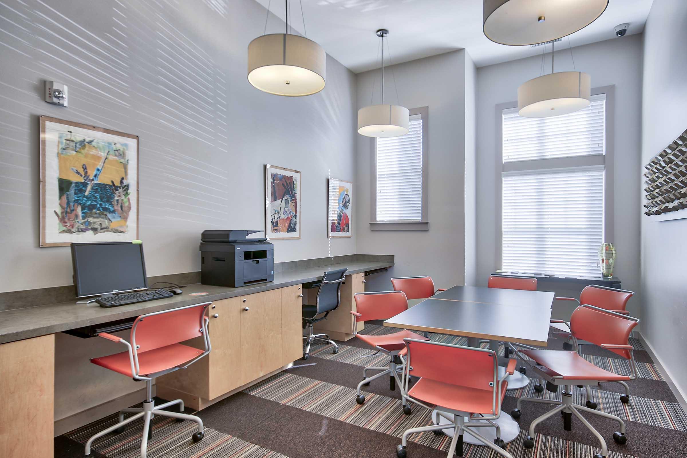 A modern conference room featuring a large table surrounded by red chairs, a printer, and a computer setup. The walls are adorned with framed artwork, and there are large windows letting in natural light, complemented by stylish pendant lights above. The flooring has a mix of carpet patterns.