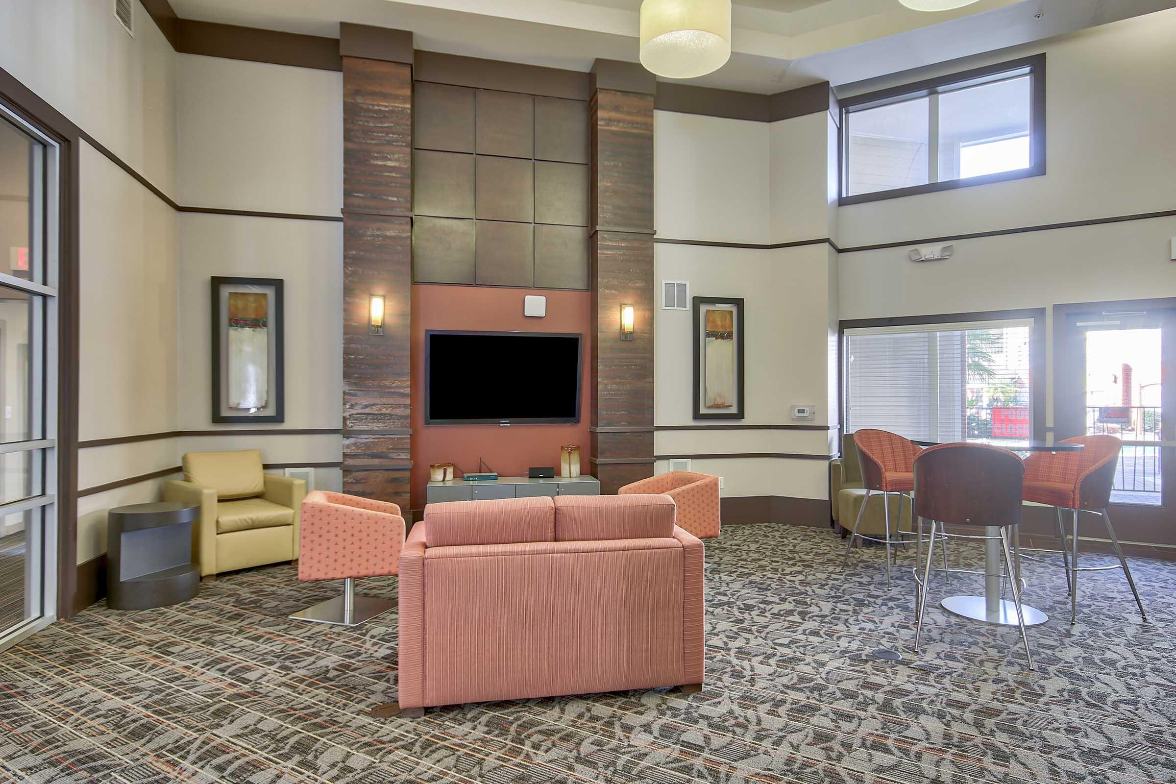 A modern lobby area featuring a cozy seating arrangement with a peach-colored sofa and armchairs. There's a large flat-screen TV mounted on the wall, and large windows provide natural light. The floor has a patterned carpet, and stylish light fixtures hang from the ceiling, creating a welcoming atmosphere.