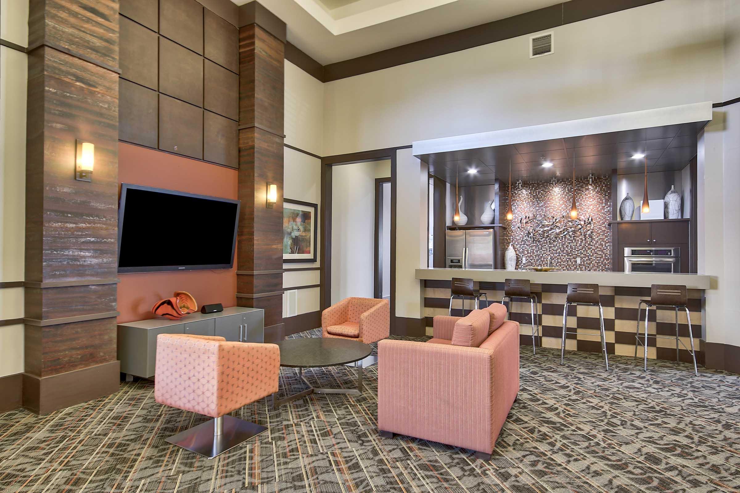 A modern lounge area featuring a contemporary design with two pink armchairs, a round black coffee table, a large flat-screen TV mounted on the wall, and a sleek kitchen bar in the background with stools. The decor includes a mix of textures and colors, creating a welcoming atmosphere.
