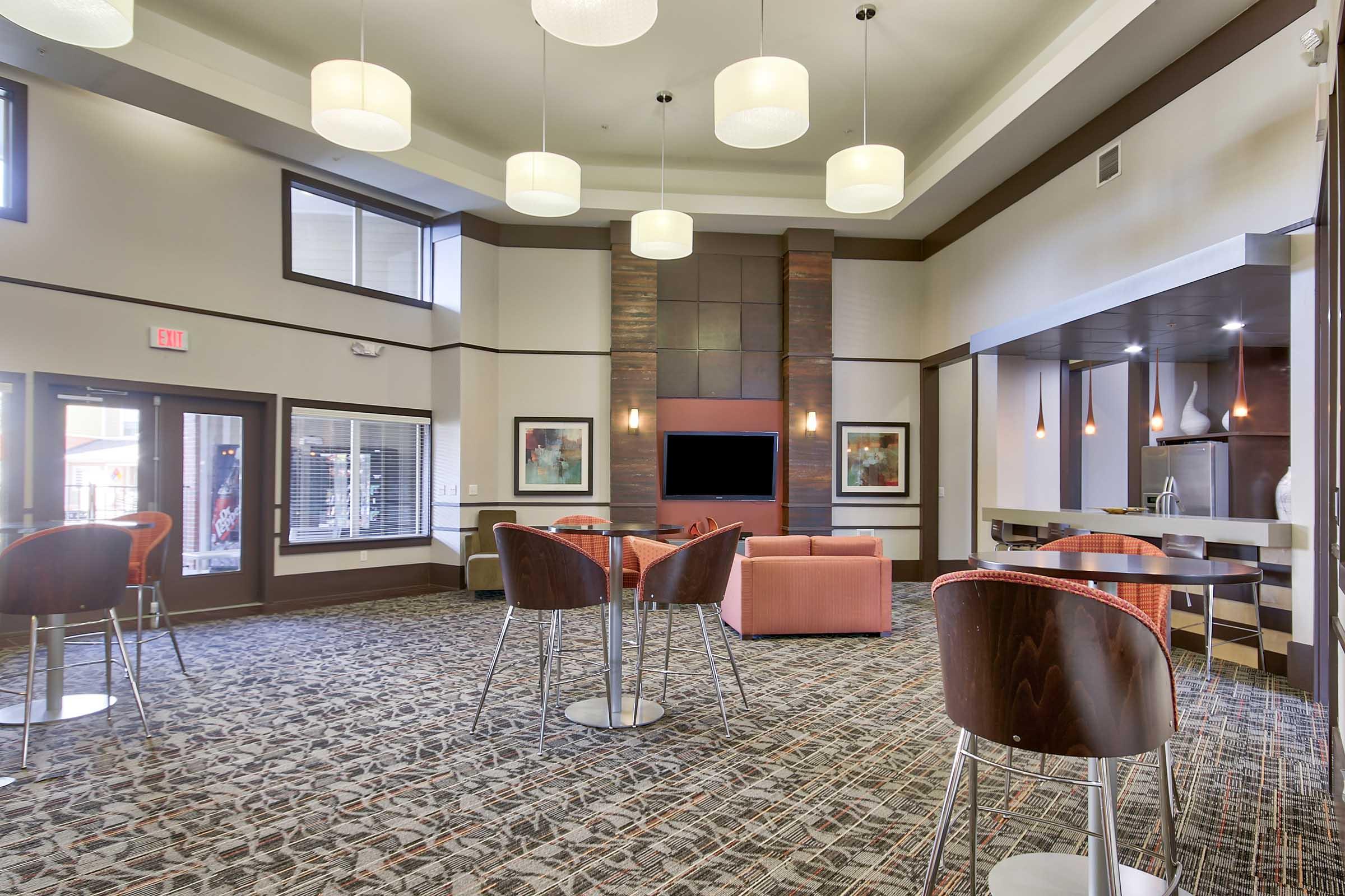 A modern lounge area with a carpeted floor, featuring stylish furniture including brown chairs and orange sofas. The space has large windows, decorative wall art, and a central flat-screen TV, with ambient lighting from pendant lamps hanging from the ceiling.
