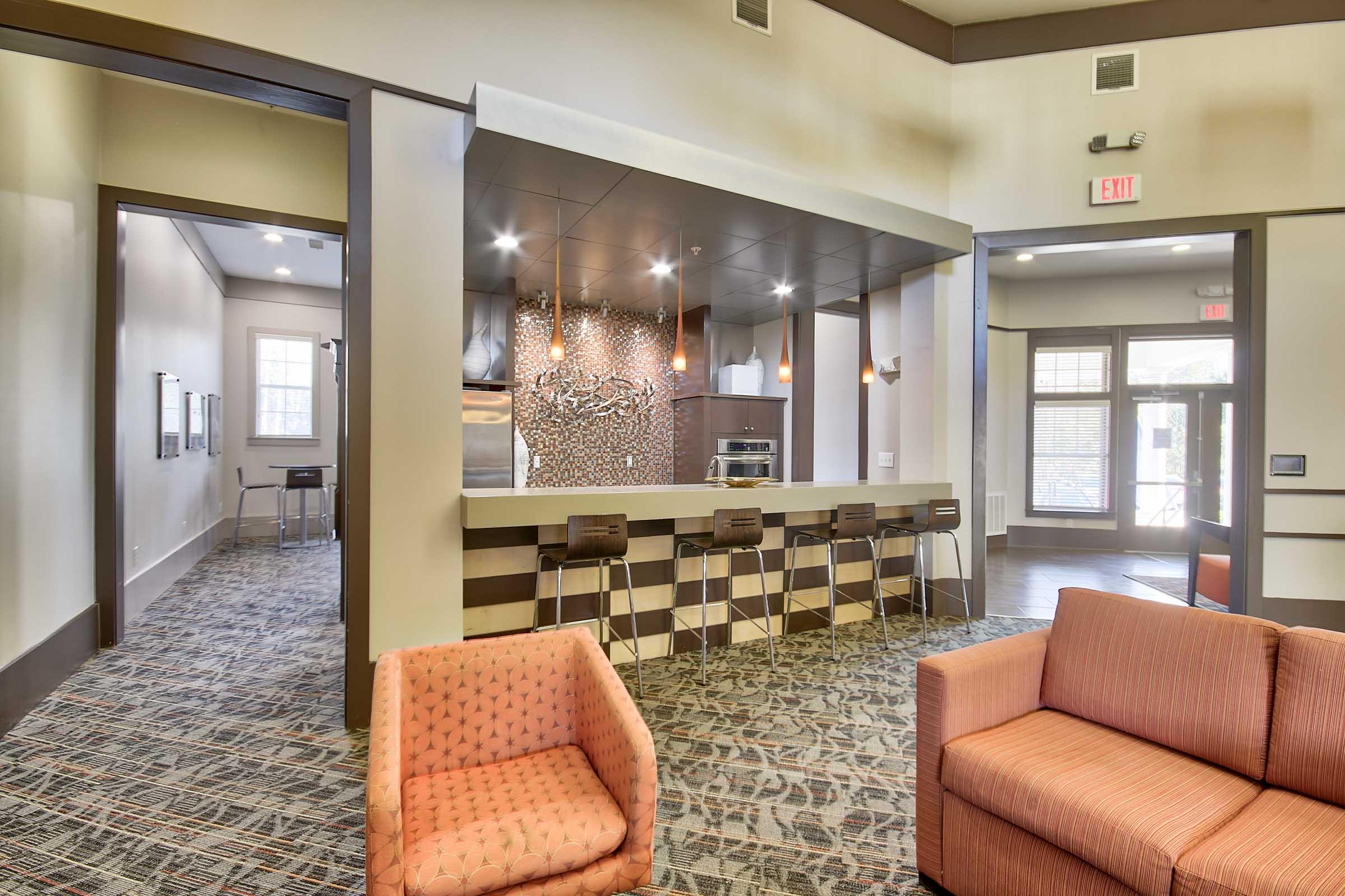 A modern communal kitchen area with a sleek bar featuring high stools. The walls are adorned with patterned tiles, and a cozy seating area with two stylish armchairs is visible in the foreground. Natural light floods the space through large windows, creating an inviting atmosphere.