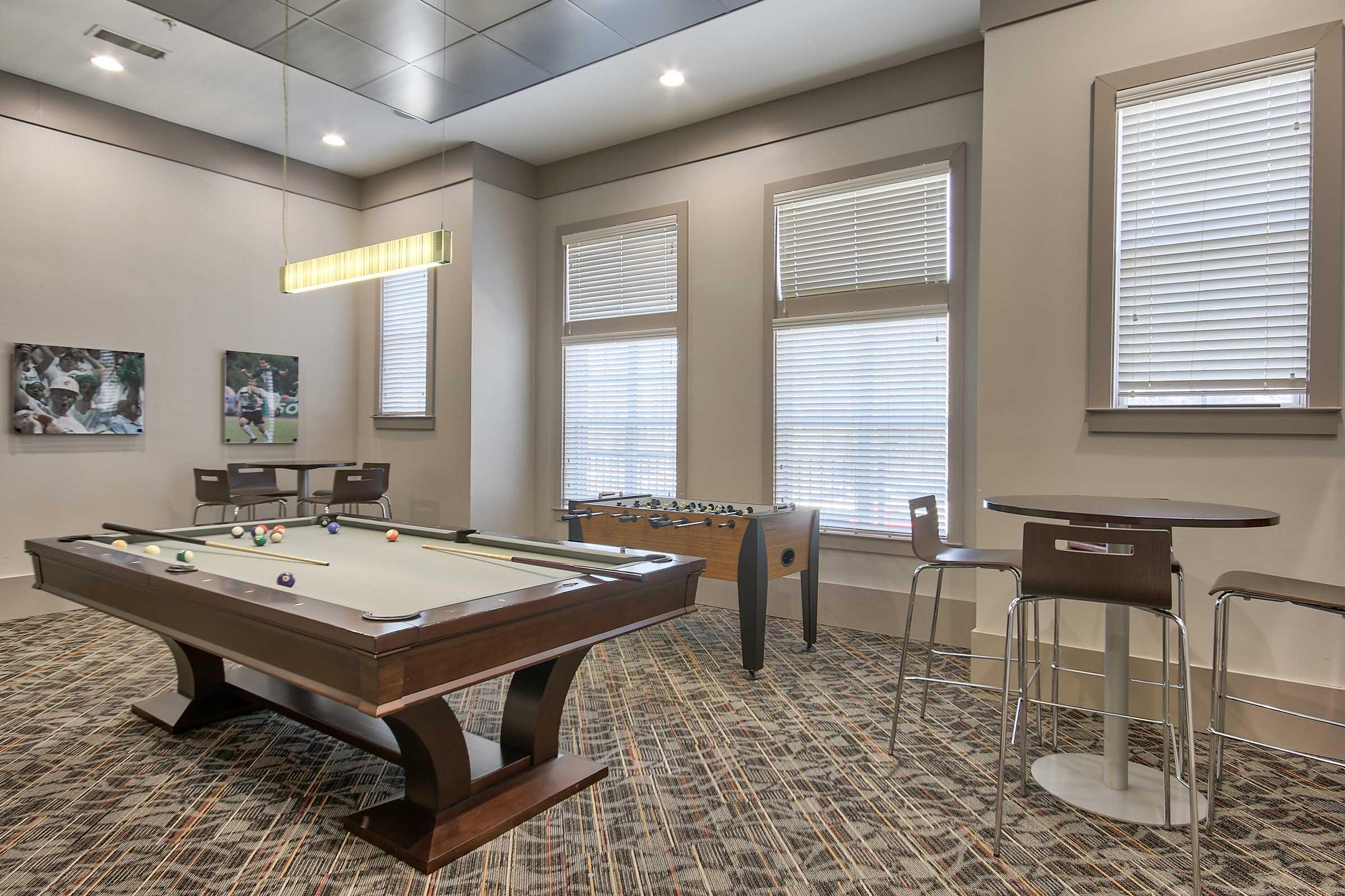 A cozy recreation room featuring a pool table, a foosball table, and a small bar table with chairs. Large windows allow natural light to brighten the space, which has contemporary decor and a carpeted floor. Two wall-mounted pictures add to the inviting atmosphere.