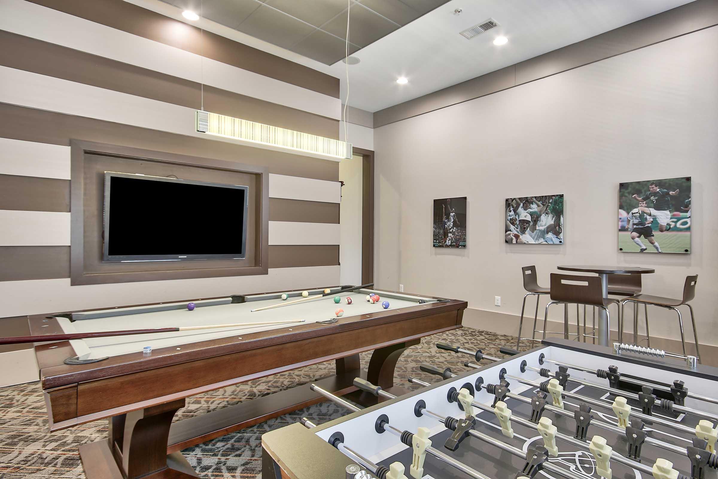 A modern recreational room featuring a pool table and a foosball table. The walls are decorated with striped designs, and there is a flat-screen TV mounted on the wall. Two framed images hang on the adjacent wall, and a small table with chairs is situated nearby. The floor has a textured pattern.