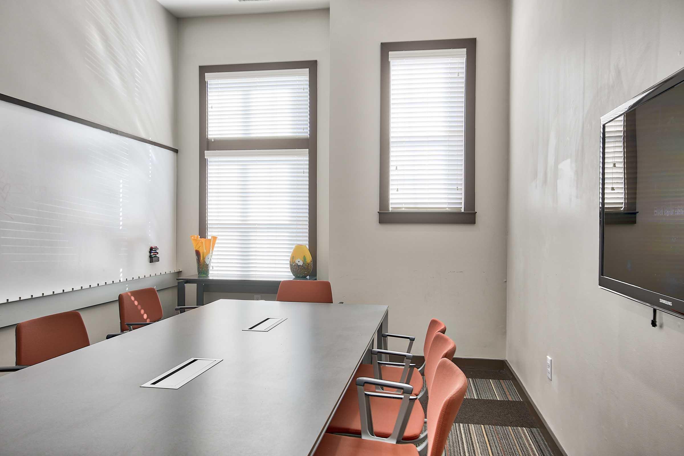 A modern conference room featuring a large gray table with multiple chairs, a whiteboard on one wall, and windows with blinds allowing natural light. A small vase with orange flowers is placed on the table, adding a touch of color to the neutral-toned room. A flat-screen TV is mounted on another wall.