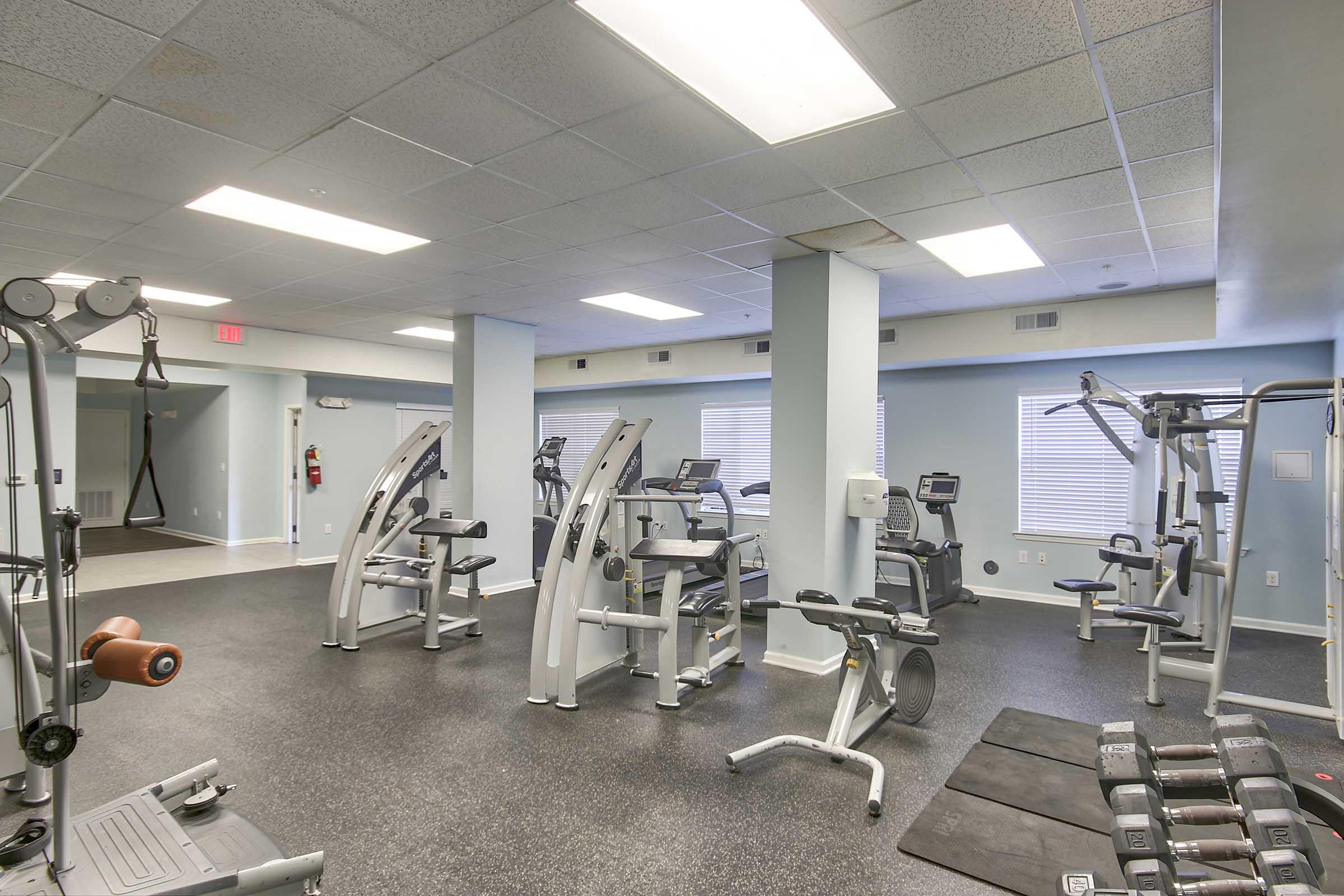 A well-lit gym interior featuring various exercise machines, including weight machines and cardio equipment. The floor is covered in dark rubber flooring, and there are large windows providing natural light. A set of free weights is visible in the foreground.