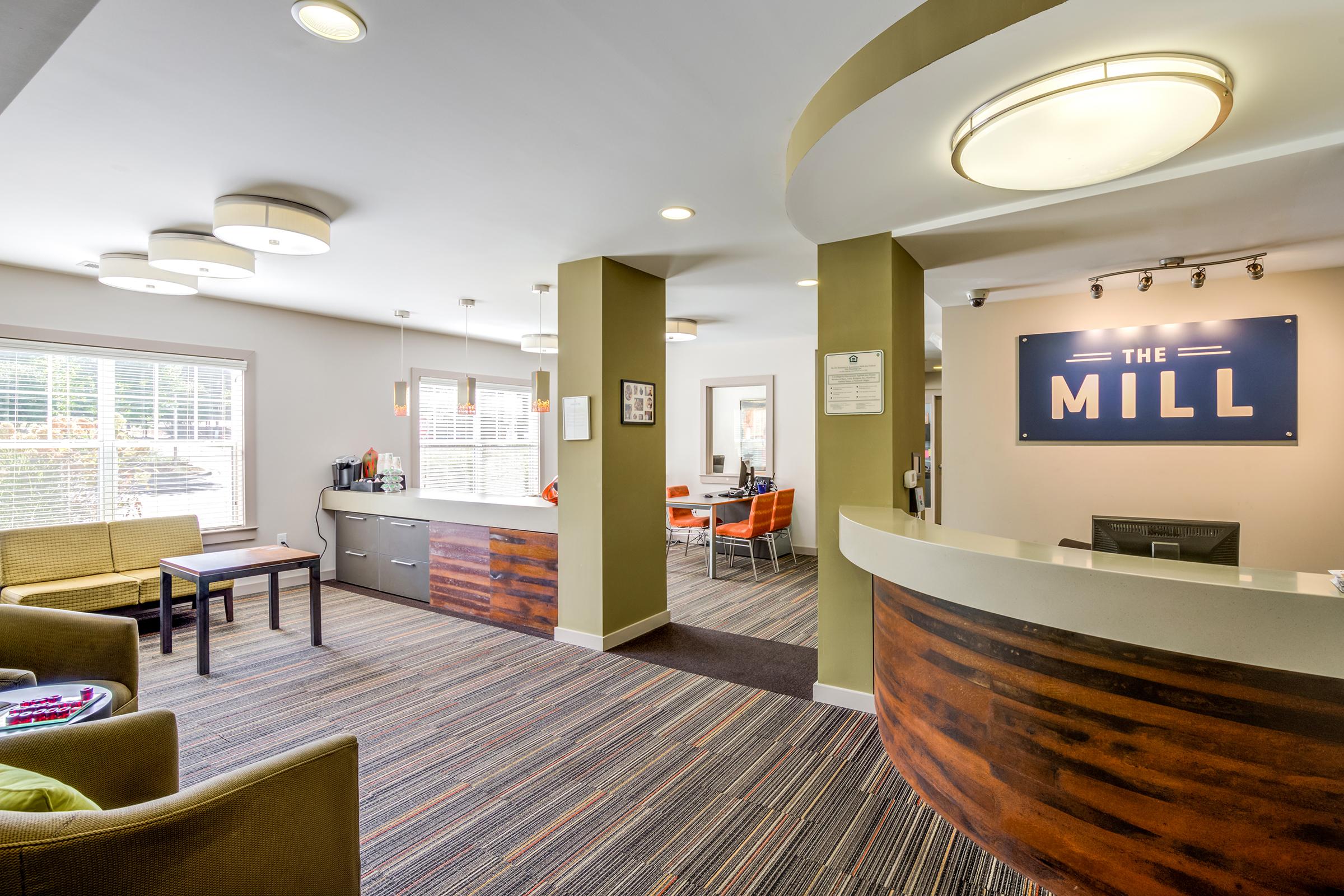Interior view of a modern reception area called "The Mill." Features include a curved reception desk, lounge seating with a coffee table, and a bright, airy atmosphere with large windows. The decor is contemporary, with a mix of neutral colors and wooden accents. Stylish lighting fixtures enhance the space.