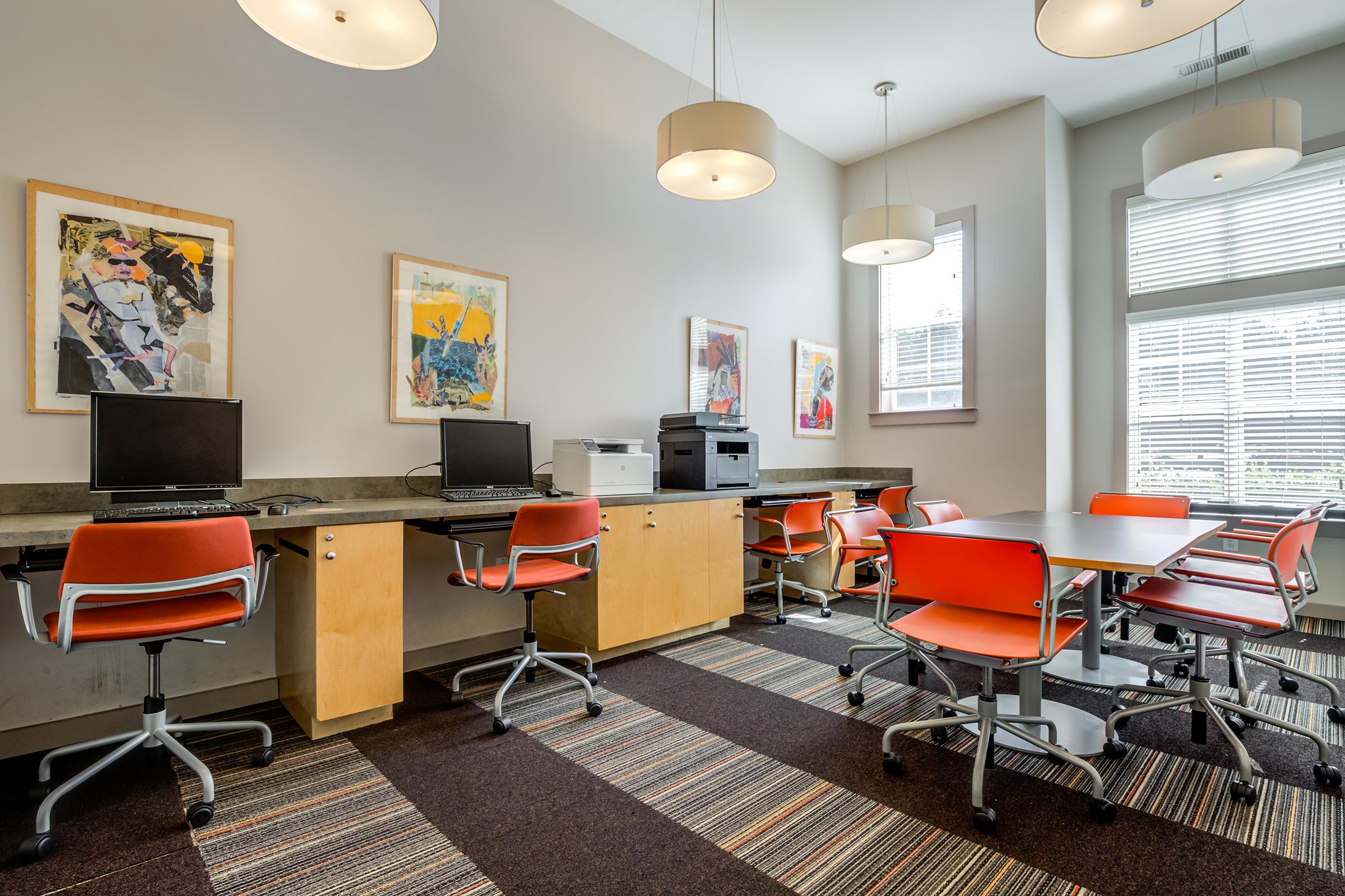 A modern workspace featuring two computers on wooden desks, a printer, and several orange swivel chairs around tables. The walls are adorned with colorful artwork, and large windows provide natural light. The flooring features a patterned carpet, creating a bright and inviting environment for work or study.