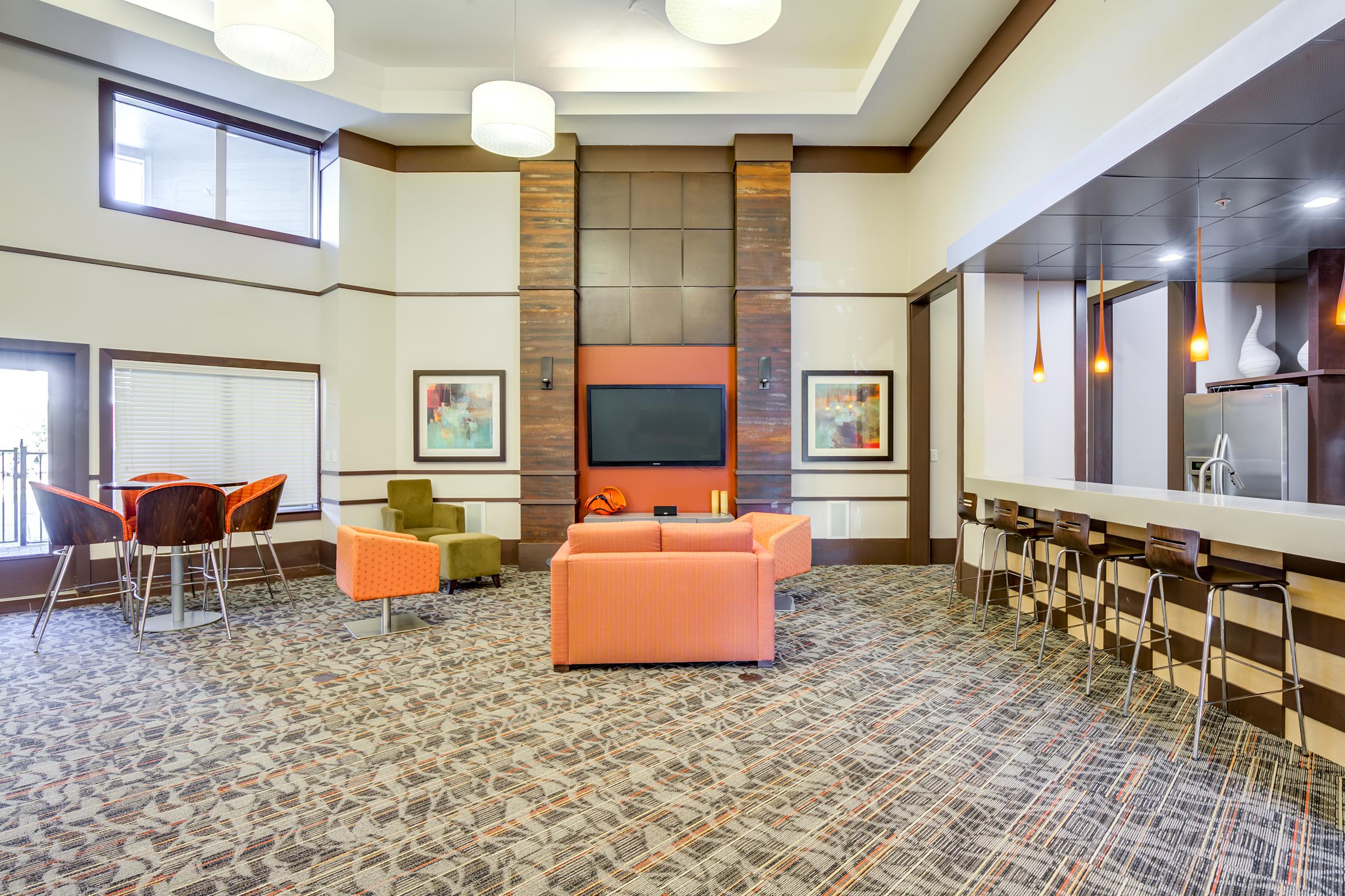 Modern interior of a stylish lounge area featuring an orange sofa, green accent chairs, a dining table with bar stools, and a large flat-screen TV mounted on a wooden panel. The space has bright lighting and a patterned carpet, creating a welcoming atmosphere.