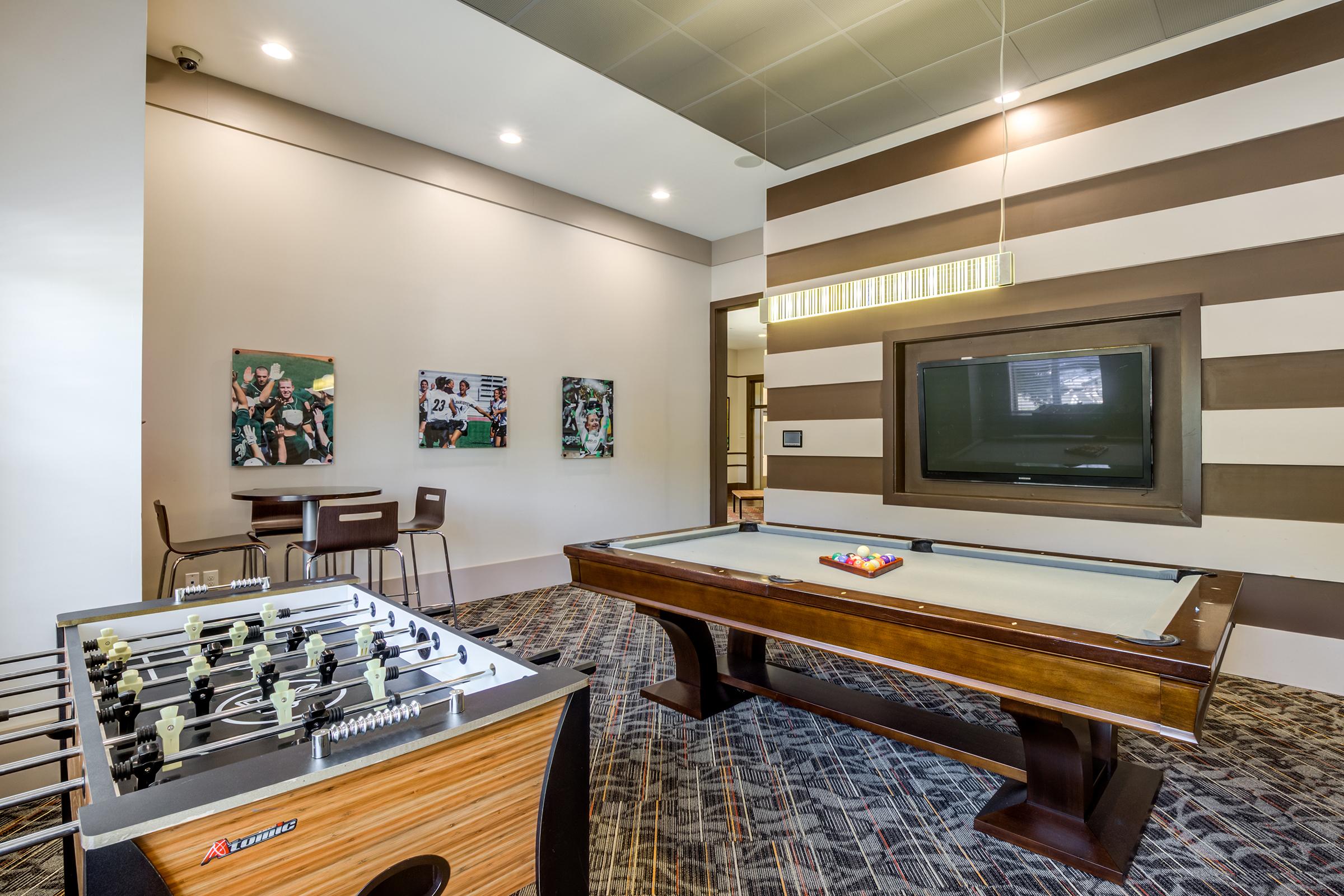 An indoor recreational area featuring a foosball table and a pool table. The walls are decorated with colorful photographs, and there’s a large flat-screen TV mounted on the wall. The space has modern lighting and a contemporary design, creating an inviting atmosphere for leisure activities.