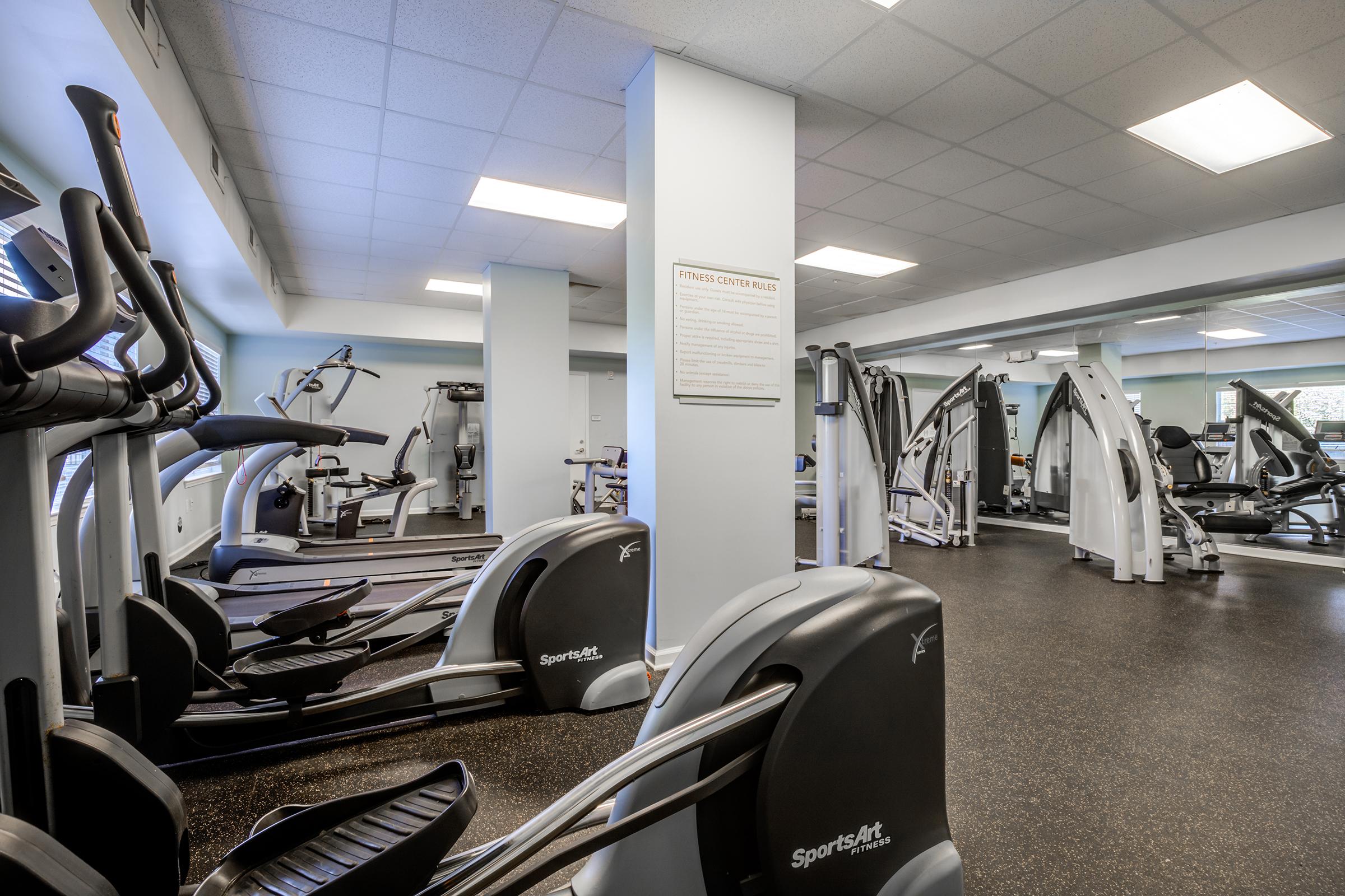 A well-lit gym interior featuring various exercise equipment, including elliptical machines, weight machines, and treadmills. The space has a polished floor and mirrors on the walls, providing a clean and modern environment for workouts. A fitness guidelines poster is visible on the wall.