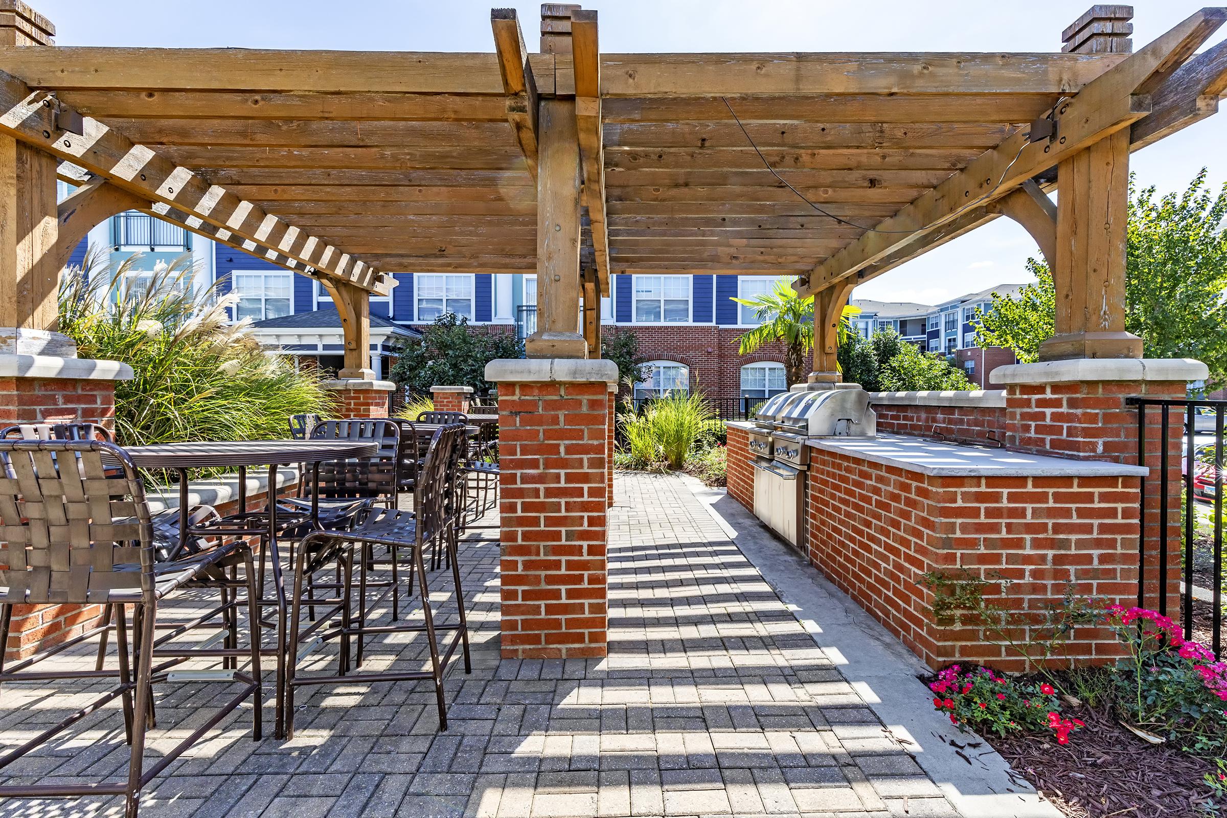 Outdoor patio area featuring a wooden pergola, brick columns, and metal bar stools around a table. An outdoor grilling station is visible on one side, surrounded by lush greenery and flowering plants. The scene is bright and inviting, with a clean paved walkway leading through the space.