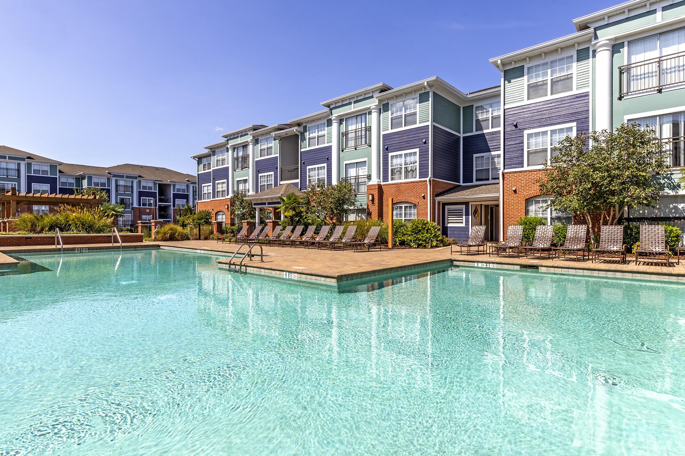 Swimming pool surrounded by lounge chairs and landscaped greenery, with modern apartment buildings in the background under a clear blue sky.