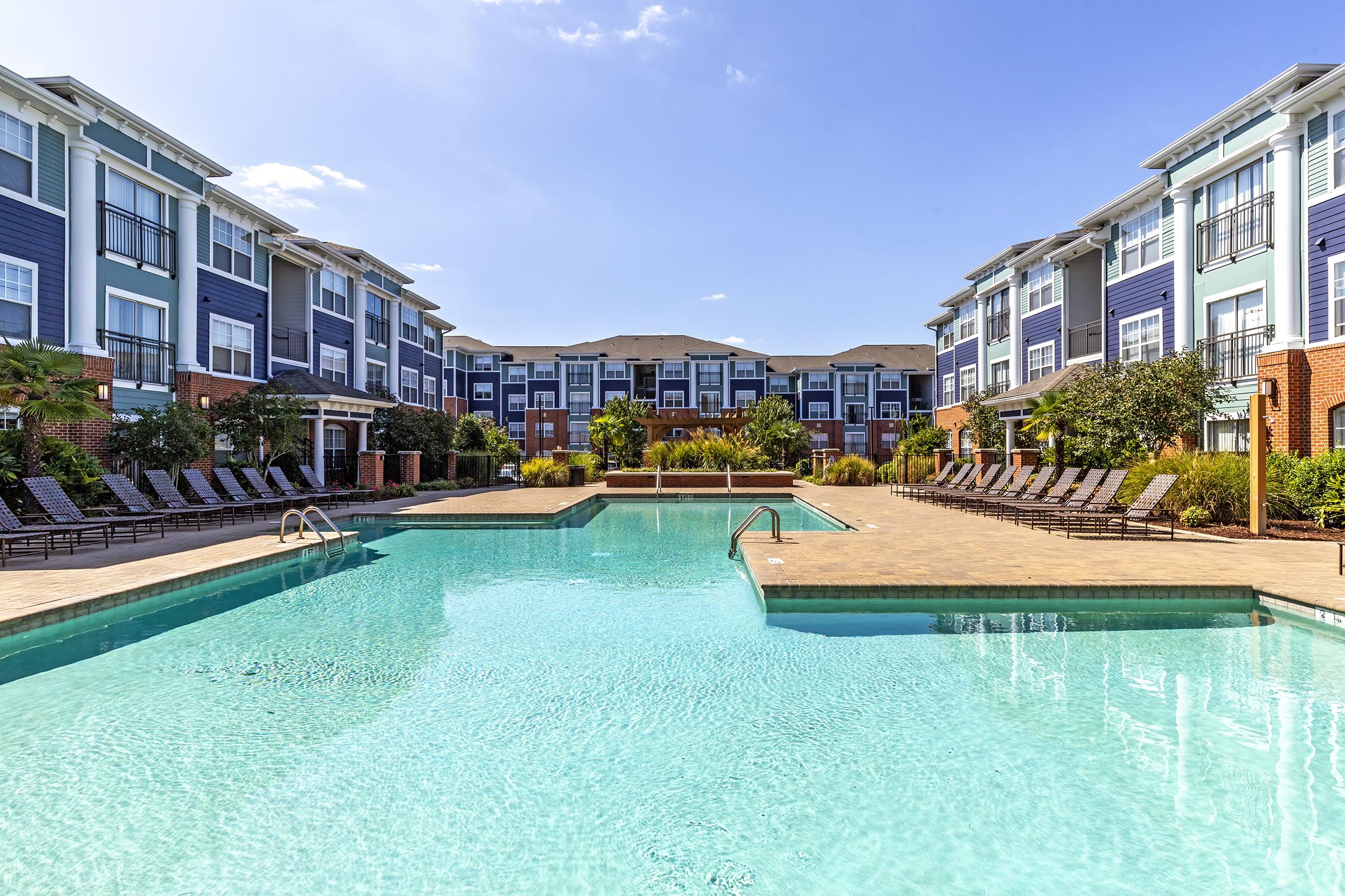 A clear pool surrounded by lounge chairs, with well-maintained landscaping and modern apartment buildings in the background under a sunny blue sky. The scene conveys a serene and inviting atmosphere for leisure and relaxation.