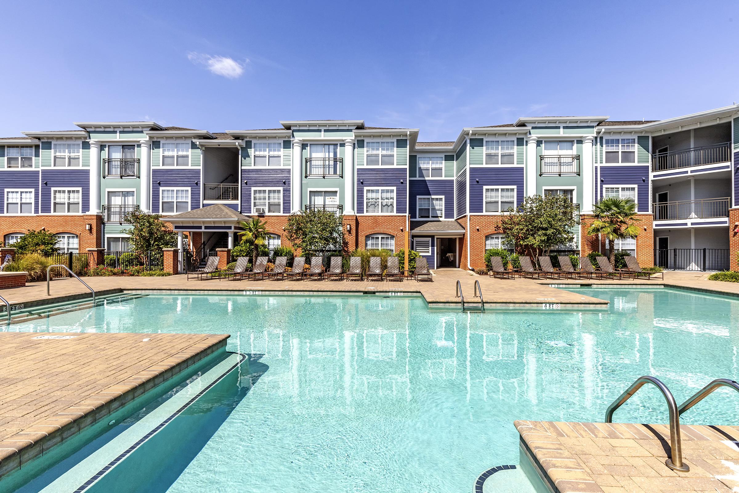 A modern apartment complex featuring a large swimming pool surrounded by lounge chairs. The building has a mix of blue and brick exteriors, with palm trees and landscaping. Clear blue sky enhances the outdoor setting, creating a welcoming atmosphere for residents and visitors.