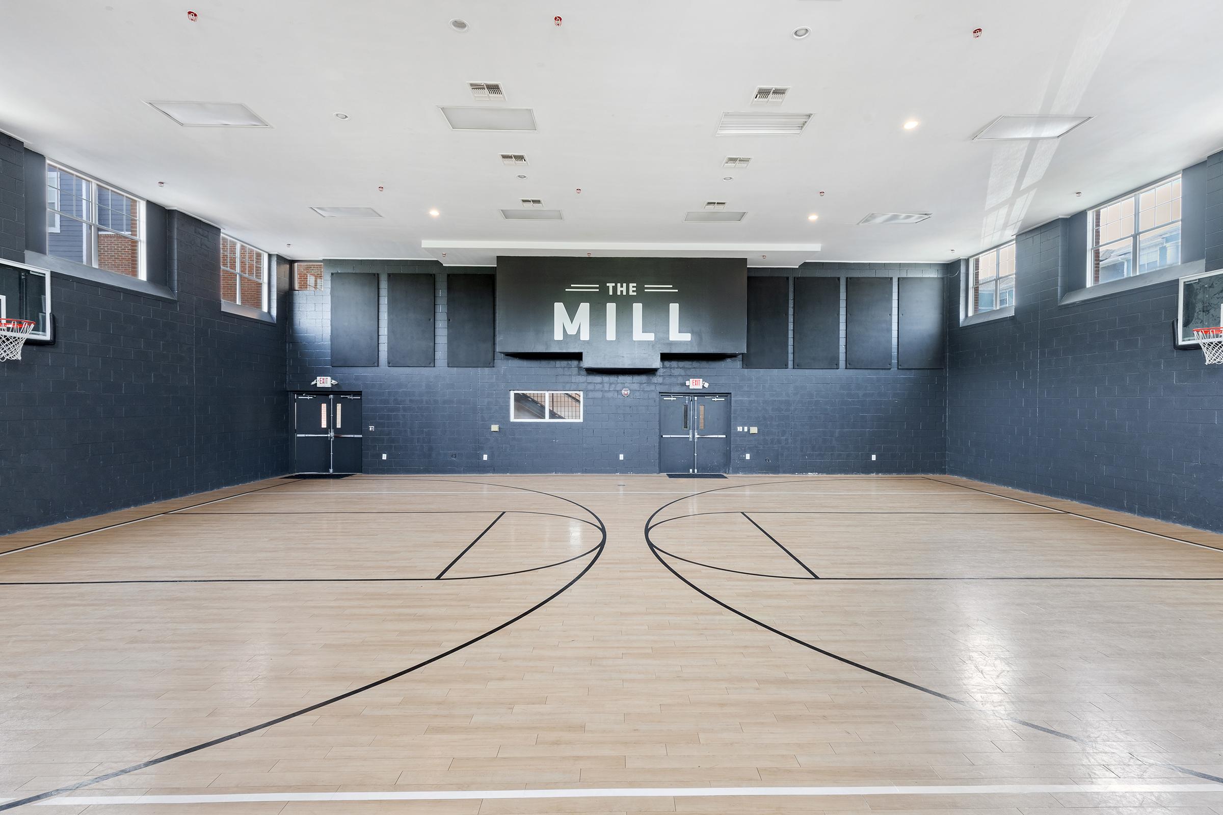 Interior of a basketball gymnasium featuring a polished wooden floor, two basketball hoops at each end, and a wall with the word "THE MILL" prominently displayed. The space has large windows providing natural light and a high ceiling, creating an open and inviting atmosphere for sports and activities.