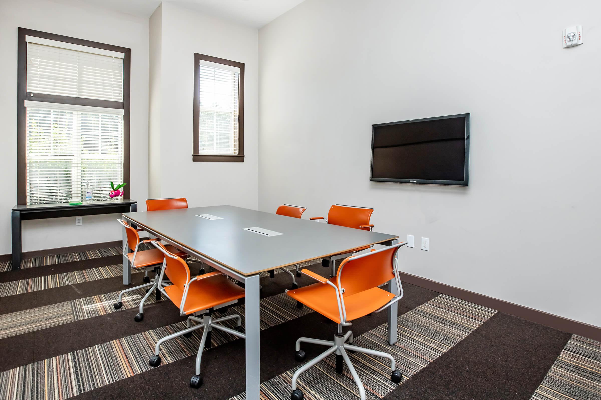 A modern conference room featuring a gray rectangular table surrounded by six orange chairs. There is a large wall-mounted TV and a window with natural light, along with a small decorative plant on a windowsill. The floor has a striped carpet design with varying colors.
