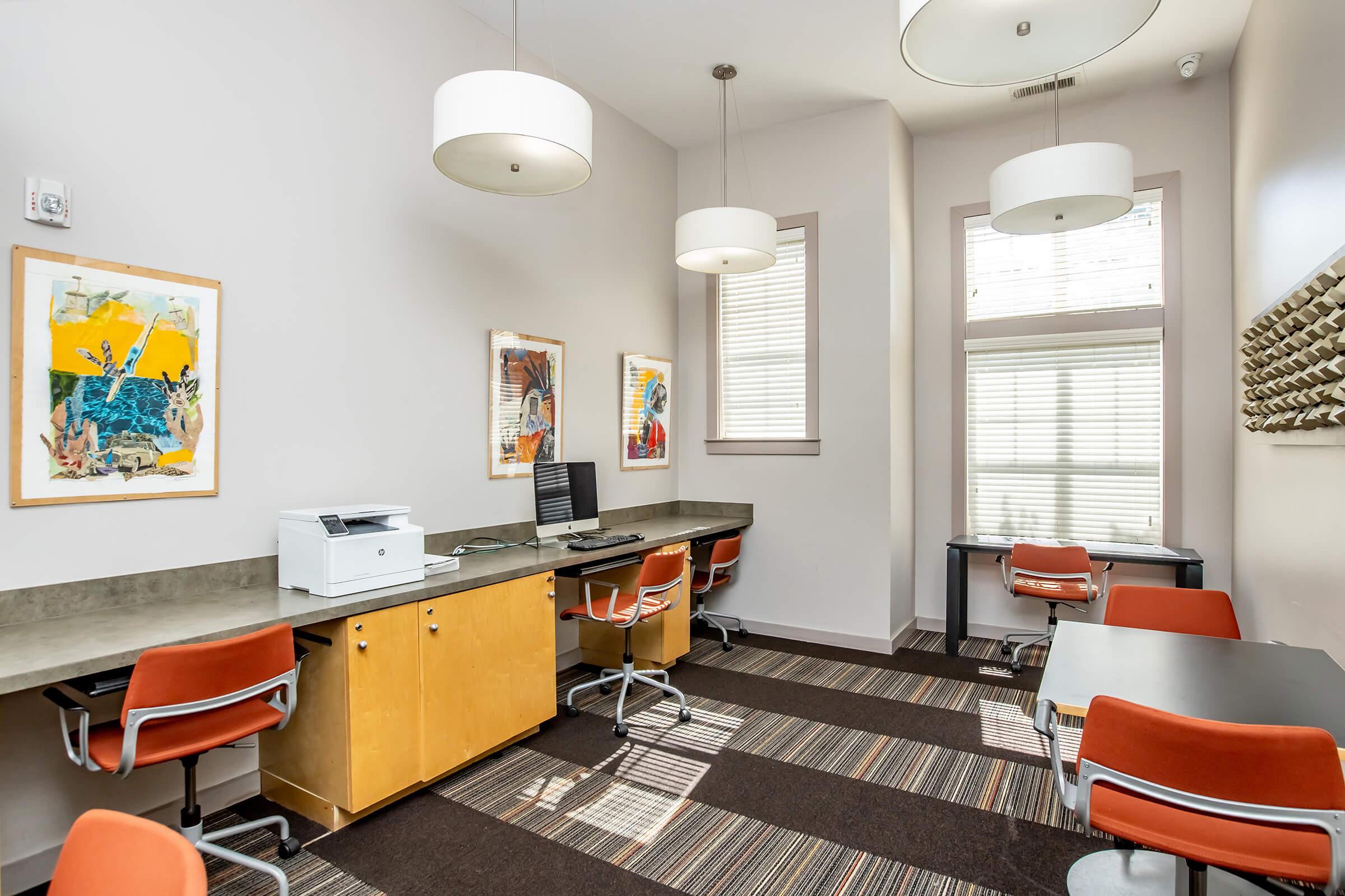 A modern office space featuring two workstations with computers, a printer, and a light-colored desk. The room has large windows with natural light, stylish overhead lights, and colorful artwork on the walls. The flooring has a striped pattern, and there are orange chairs providing a contemporary feel.