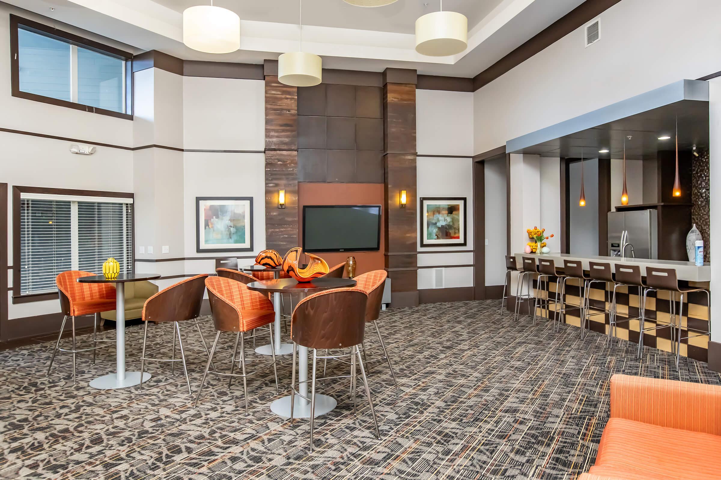 A modern lounge area featuring a seating arrangement with orange and brown chairs, high tables, and a bar counter. The space has a stylish design with artwork on the walls, large windows providing natural light, and a cozy atmosphere, ideal for relaxation or socializing.