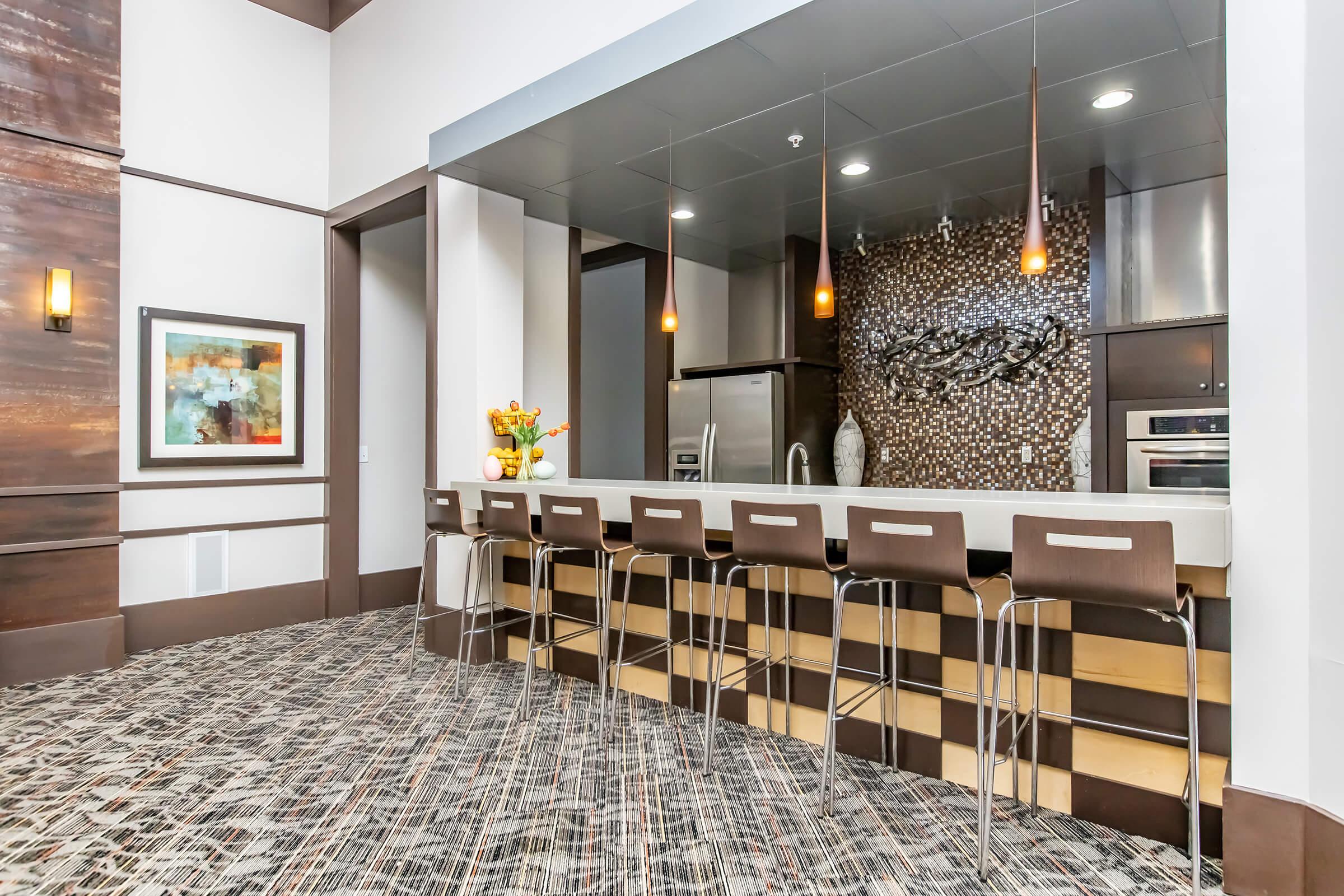 Modern kitchen featuring a stylish bar counter with several high stools, pendant lighting, and a contemporary backsplash. The space includes sleek appliances, and a decorative vase with flowers on the counter, set against a textured wall and patterned floor. Decor includes a colorful abstract painting.