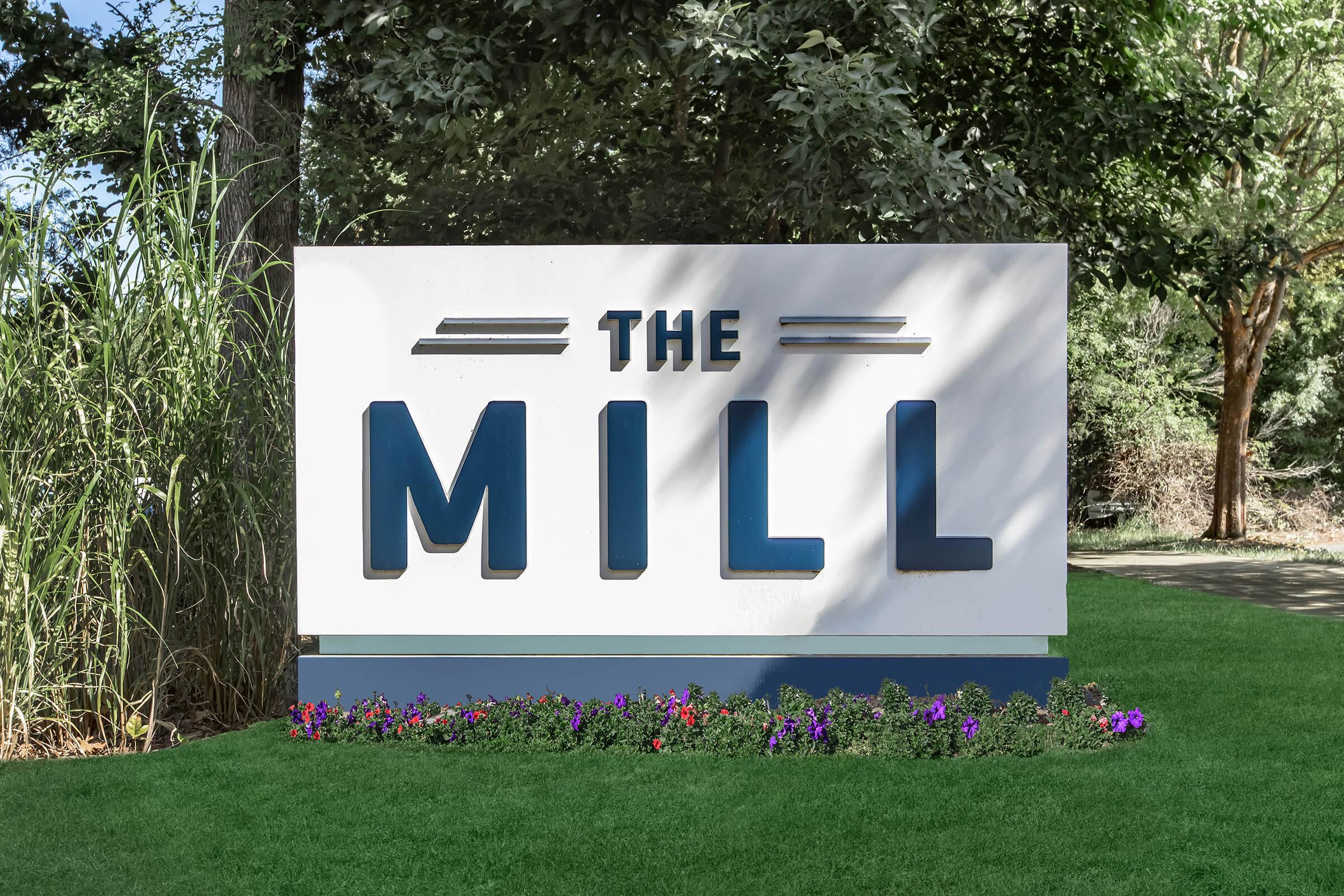 A large outdoor sign reading "THE MILL" in bold blue letters, set against a white background. The sign is placed on a grassy area with colorful flowers blooming at its base. Surrounding trees provide a natural setting.