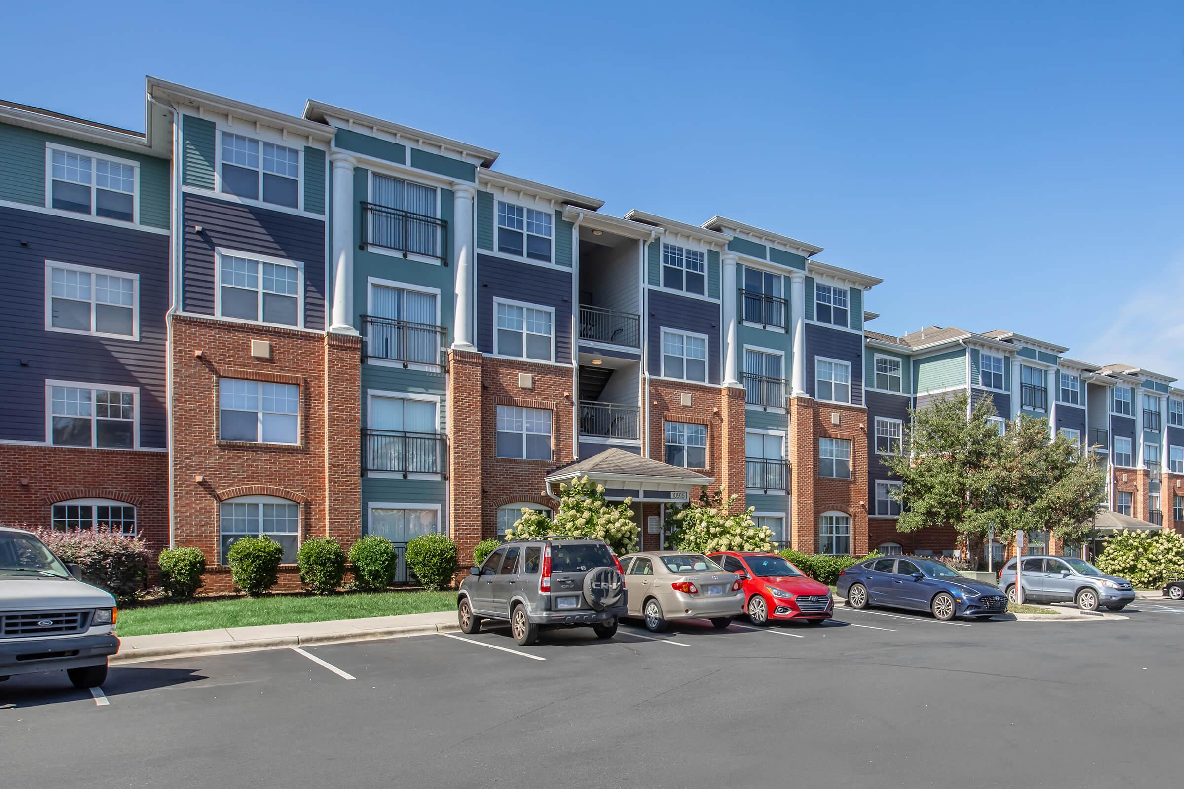 A multi-story apartment building featuring a mix of brick and colorful siding. There are landscaped areas with shrubs in front, and a parking lot with several cars parked in front of the building under clear blue skies.