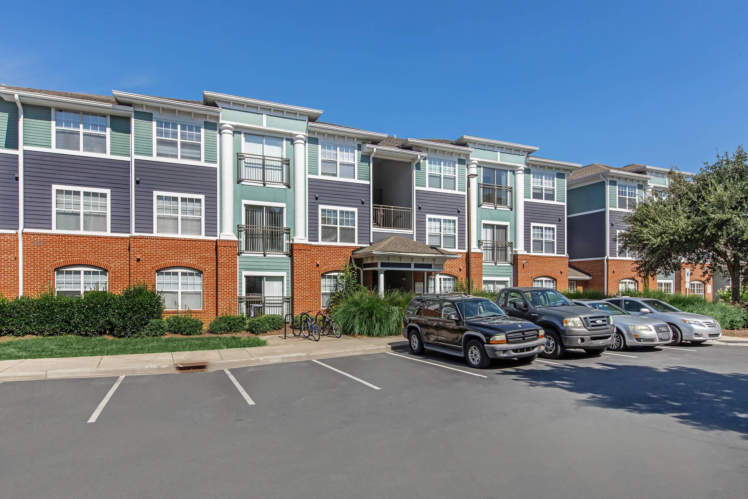 A multi-story residential apartment building with a mix of blue and brick exteriors. It features several balconies, large windows, and well-maintained landscaping with bushes. In the foreground, there are parked cars in a paved parking lot under a clear blue sky.