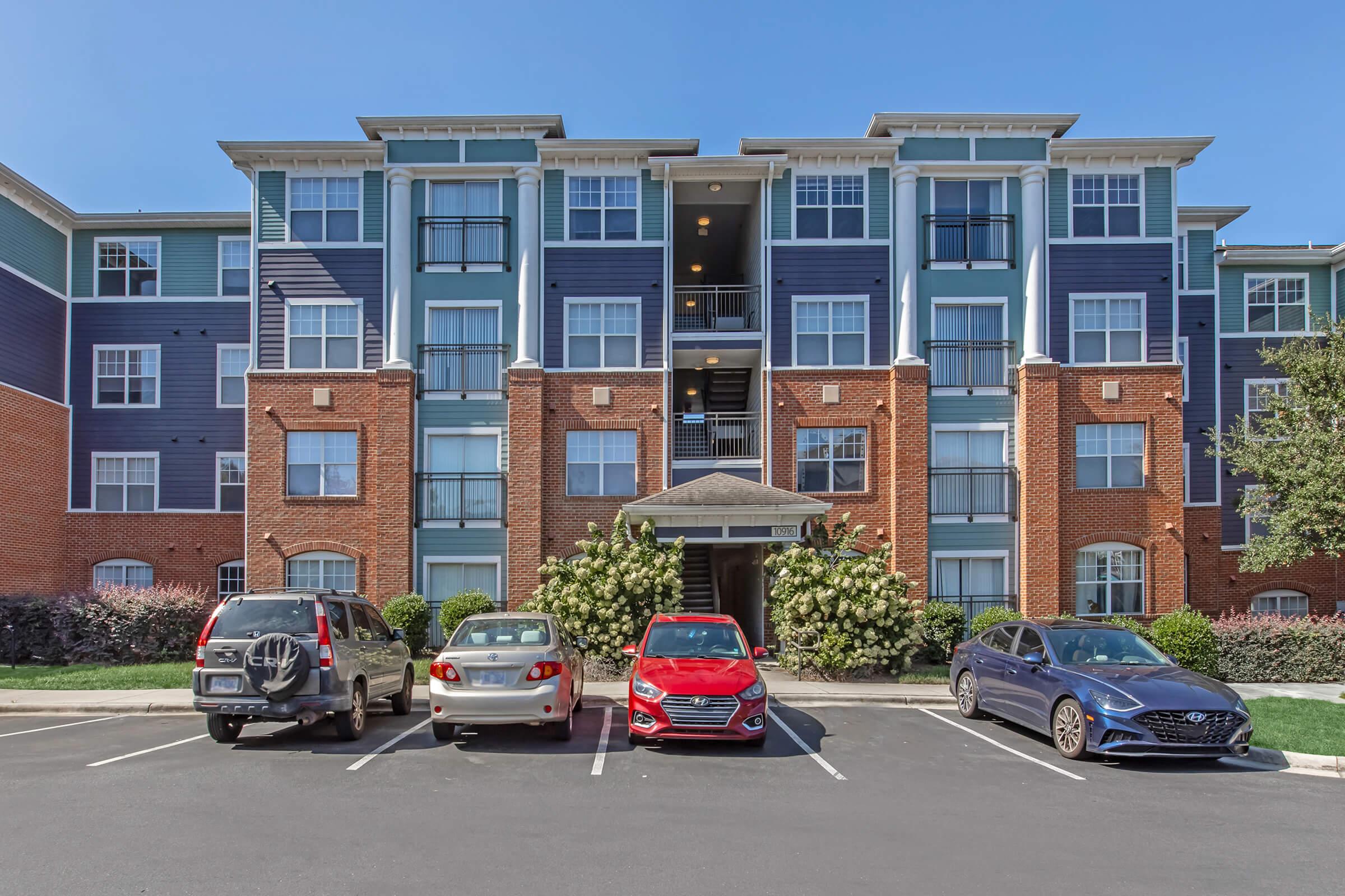 Four parked cars in front of a modern, multi-story apartment building with blue and red brick exterior. The building features balconies on each floor and is surrounded by neatly trimmed landscaping under a clear blue sky.