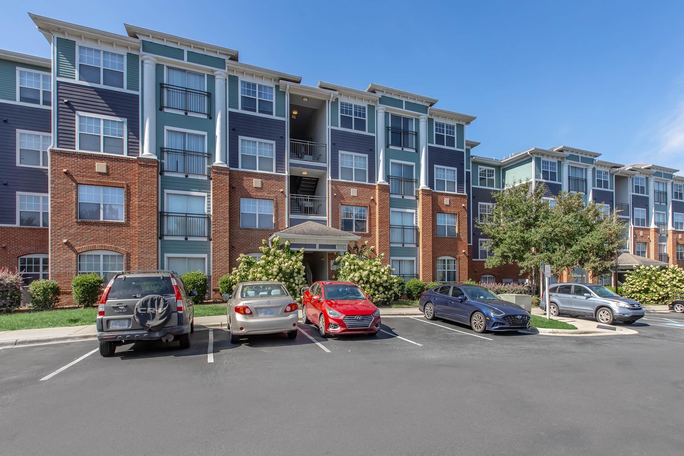 A multi-story apartment building with a mix of brick and blue siding. Several vehicles are parked in front, including a gray SUV, a silver sedan, a red car, and a dark blue car. Green shrubs and trees are planted around the parking area, with a clear blue sky overhead.