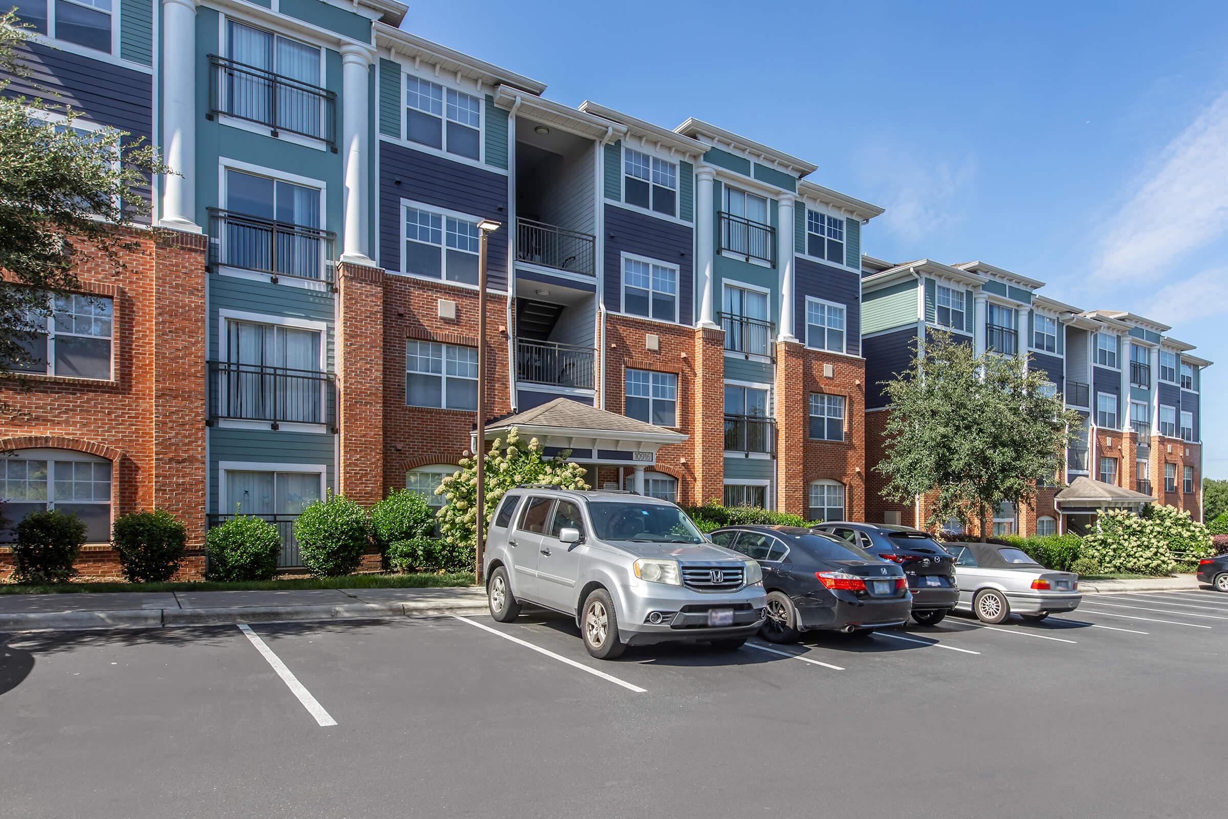 Modern apartment building with a mix of brick and blue paneling. Several parked cars are visible in the foreground. The scene is set on a sunny day with clear blue skies, showcasing well-maintained landscaping and a welcoming entrance.