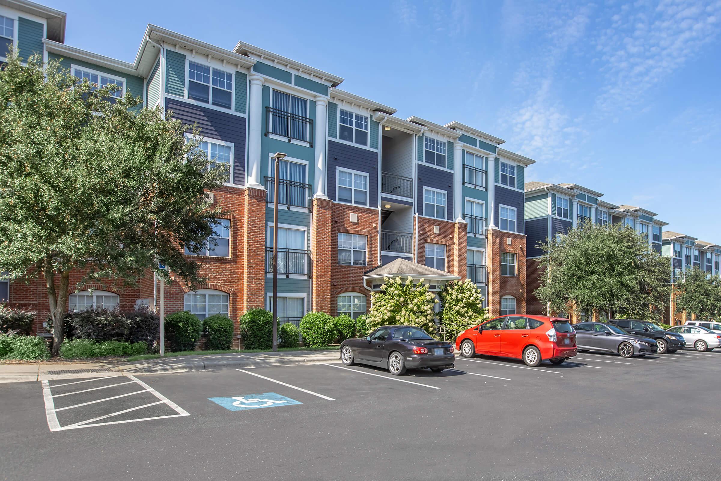 Modern three-story apartment building with blue and brick exterior, featuring landscaped greenery and parking lot. Several vehicles are parked, including a bright red car, with a few trees and bushes adding to the overall appeal of the scene. Clear blue sky in the background.