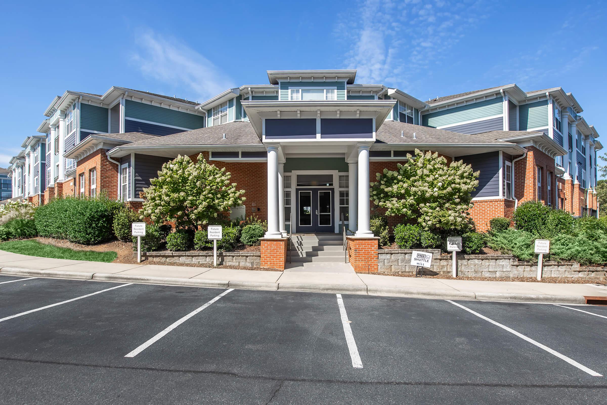 A modern brick building with multiple stories, featuring a central entrance with large windows and a covered porch. Flanking the entrance are neatly trimmed shrubs and landscaped areas. The image shows a clear blue sky and a parking area in the foreground.